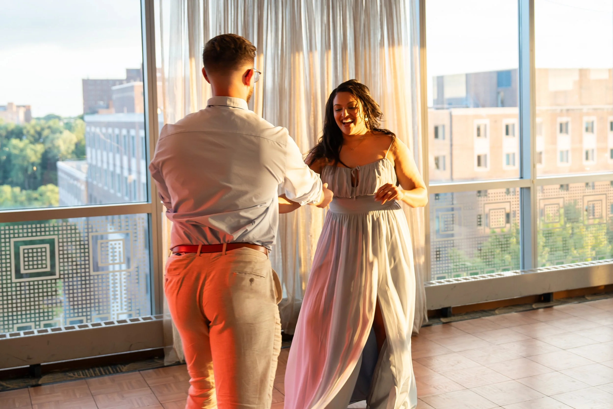 A man and woman dancing and smiling in a room with large windows during sunset, with a cityscape view in the background.