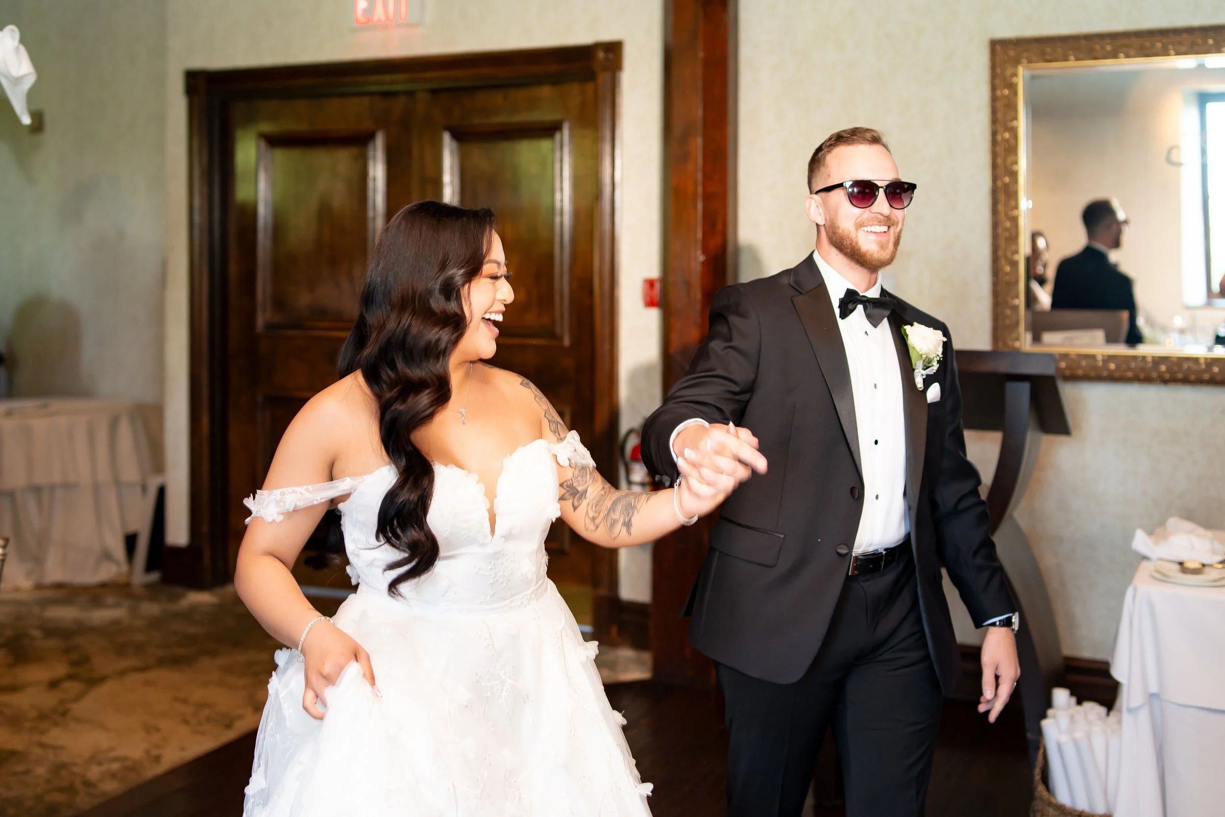 A bride and groom walking hand in hand during their wedding reception, smiling and laughing, with the bride in a white wedding dress and the groom in a black tuxedo with sunglasses.