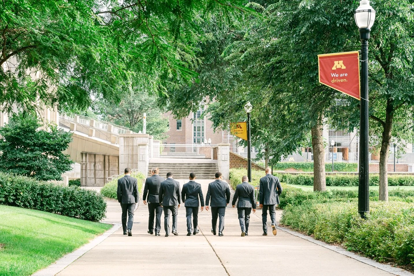 A group of eight men in business suits walking on a path through a green park area with trees and lampposts, under banners that say "We are driven".