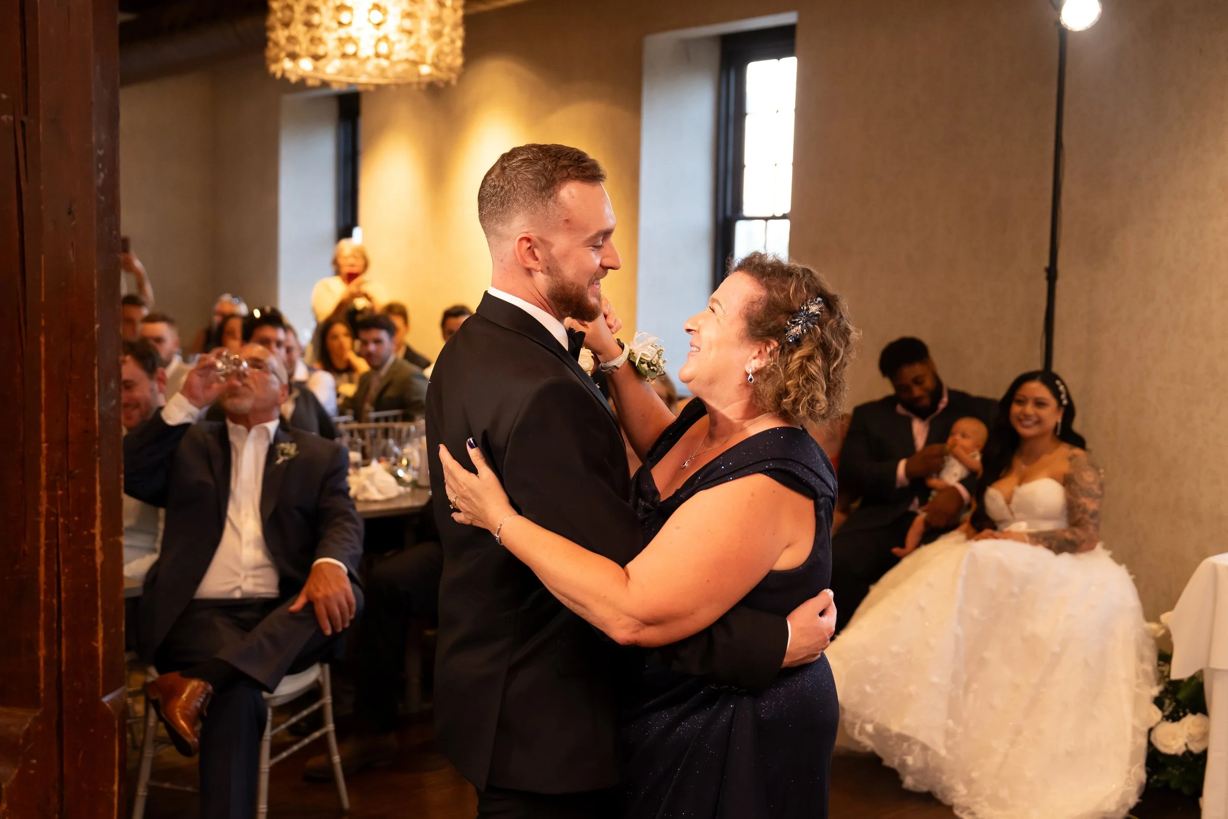 A young man in a tuxedo dancing with an older woman in a navy blue dress during a wedding reception. Guests seated at tables in the background, including a bride in a wedding gown holding a baby, and a man in a suit. The setting is an elegant indoor 