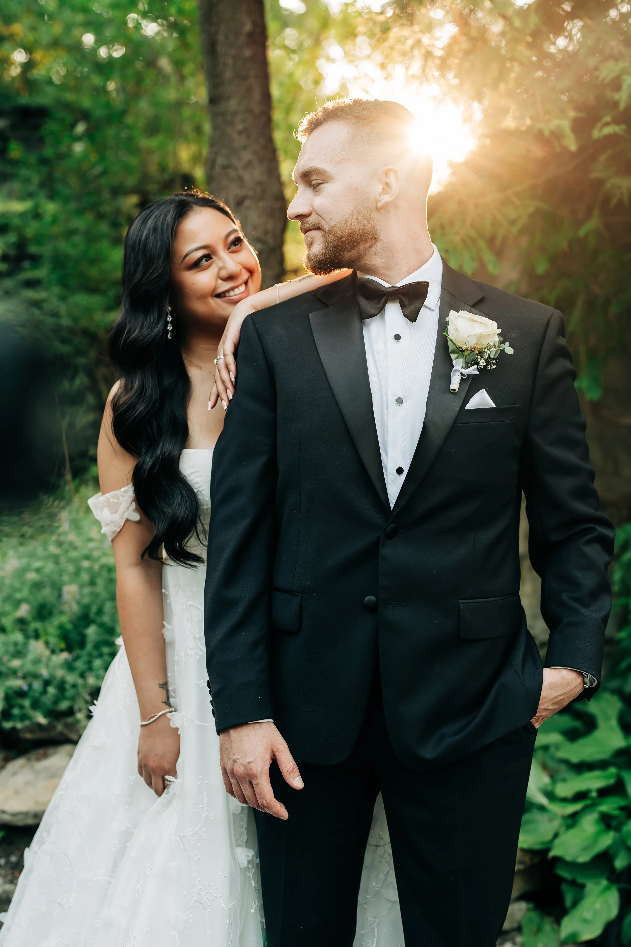 A bride and groom in wedding attire outdoors, with sunlight filtering through trees in the background.