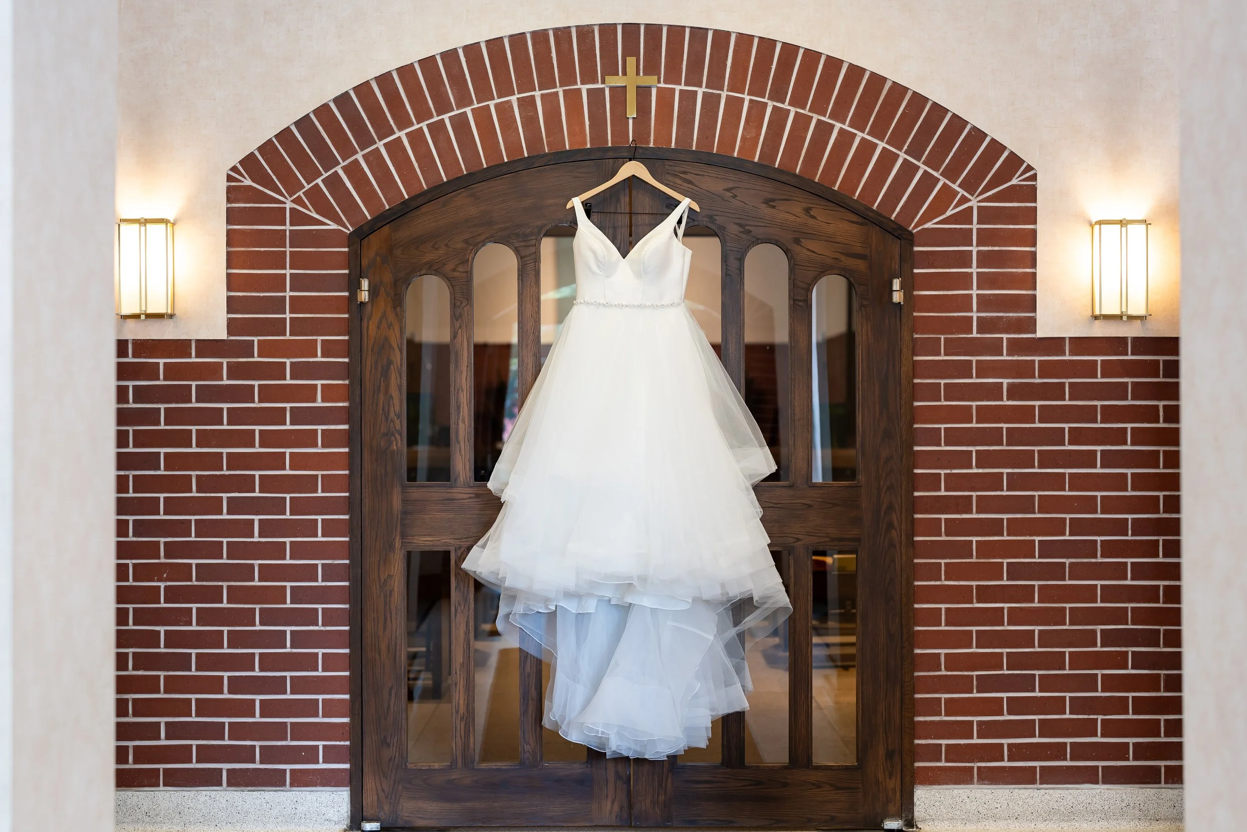 White wedding dress hanging on a hanger in front of a wooden door with glass panels, set against a brick wall with two wall sconces and a small cross above the door.