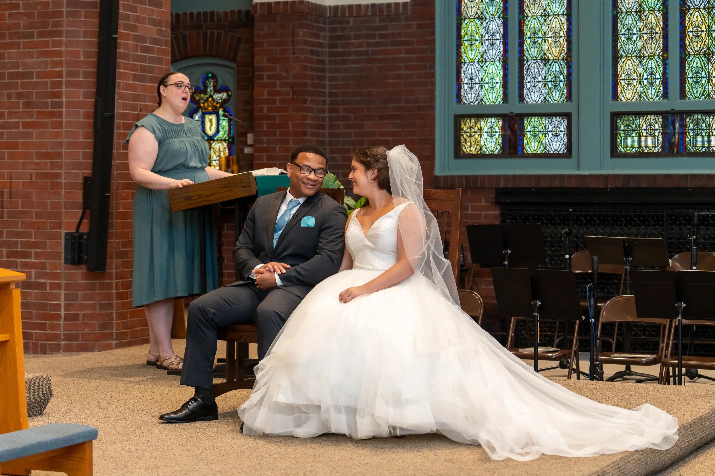 A bride and groom sit together during a wedding ceremony in a church with stained glass windows. The bride wears a white wedding dress and veil, while the groom wears a gray suit. An officiant stands behind a podium, speaking, inside a brick-walled c