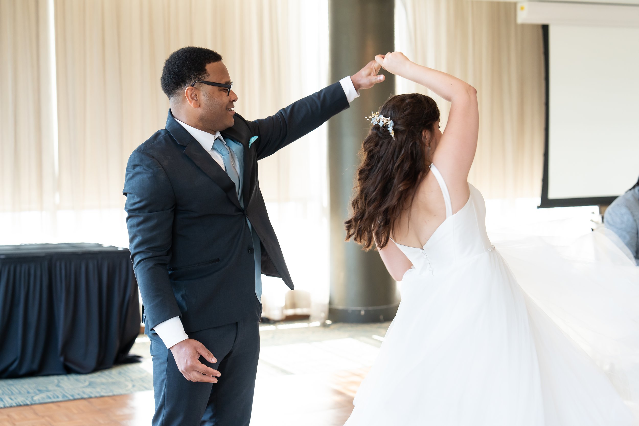 A man in a dark suit and glasses dancing with a woman in a white wedding dress, holding her hand up in a dance pose in a brightly lit room.