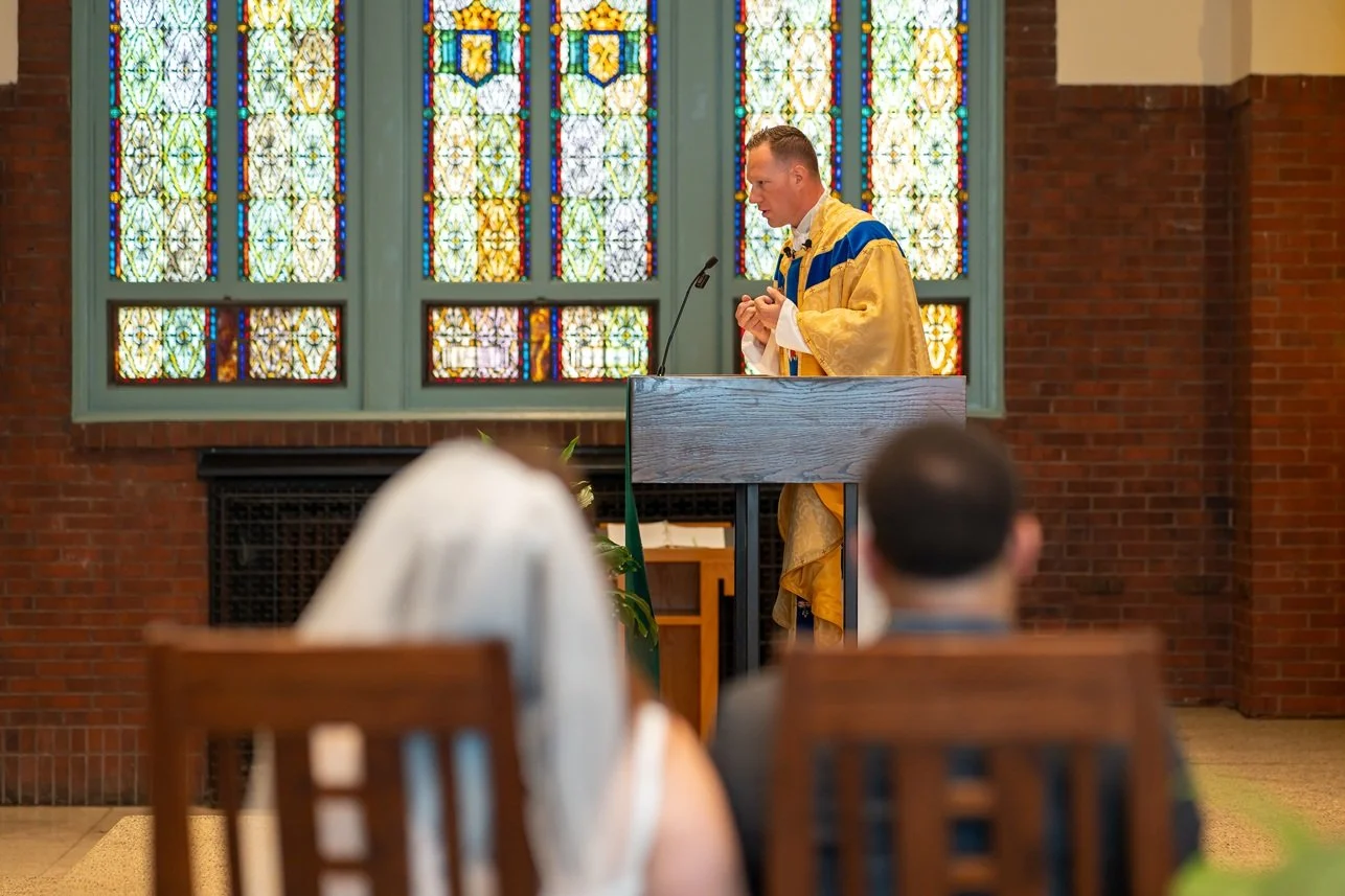 A man dressed in ceremonial robes delivering a speech or sermon at a church podium, with stained glass windows in the background and several people seated in wooden pews in the foreground.