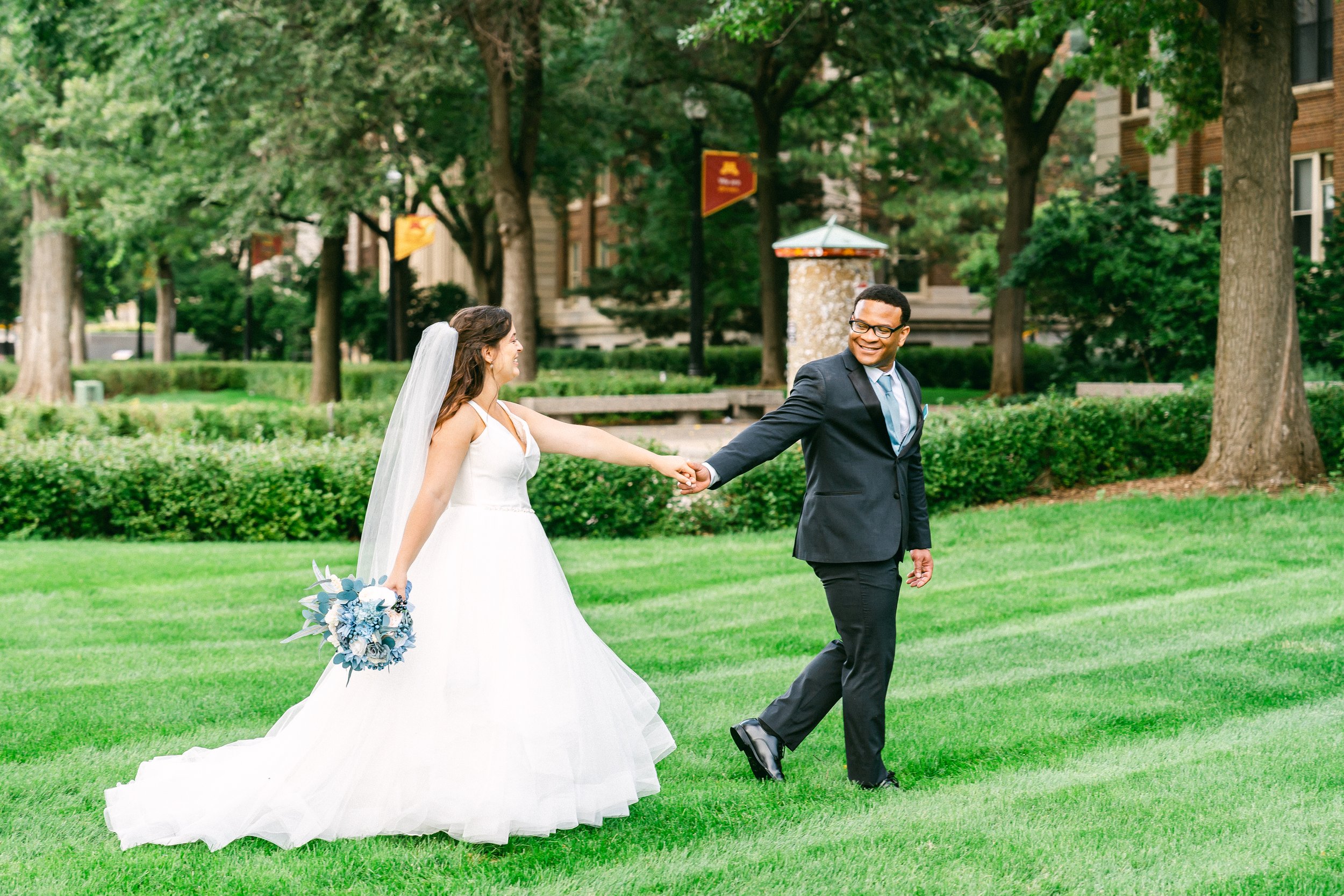 A bride in a white wedding gown holding a bouquet, and a groom in a dark suit holding her hand, walking on a grassy park with trees and buildings in the background.