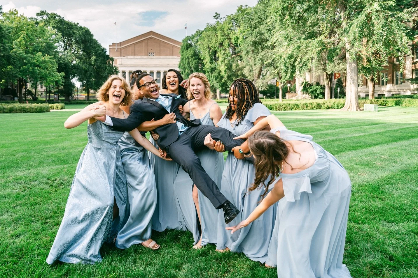 A group of women in formal dresses playfully lifting and holding a man in a suit on a lush green lawn outdoors, with a large building and trees in the background.