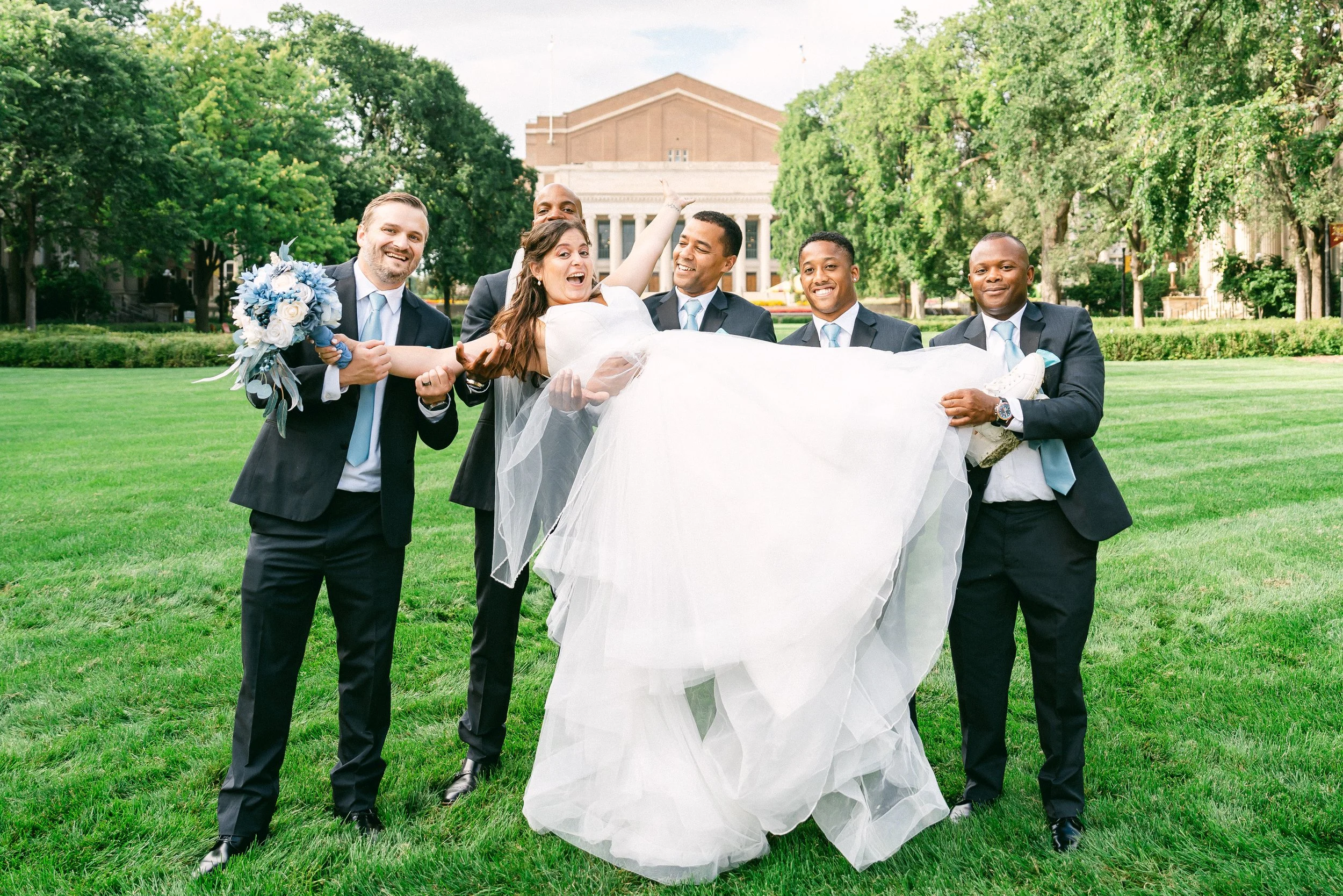 Bride in a white wedding dress being carried by five groomsmen in suits on a green lawn with trees and a large building in the background.