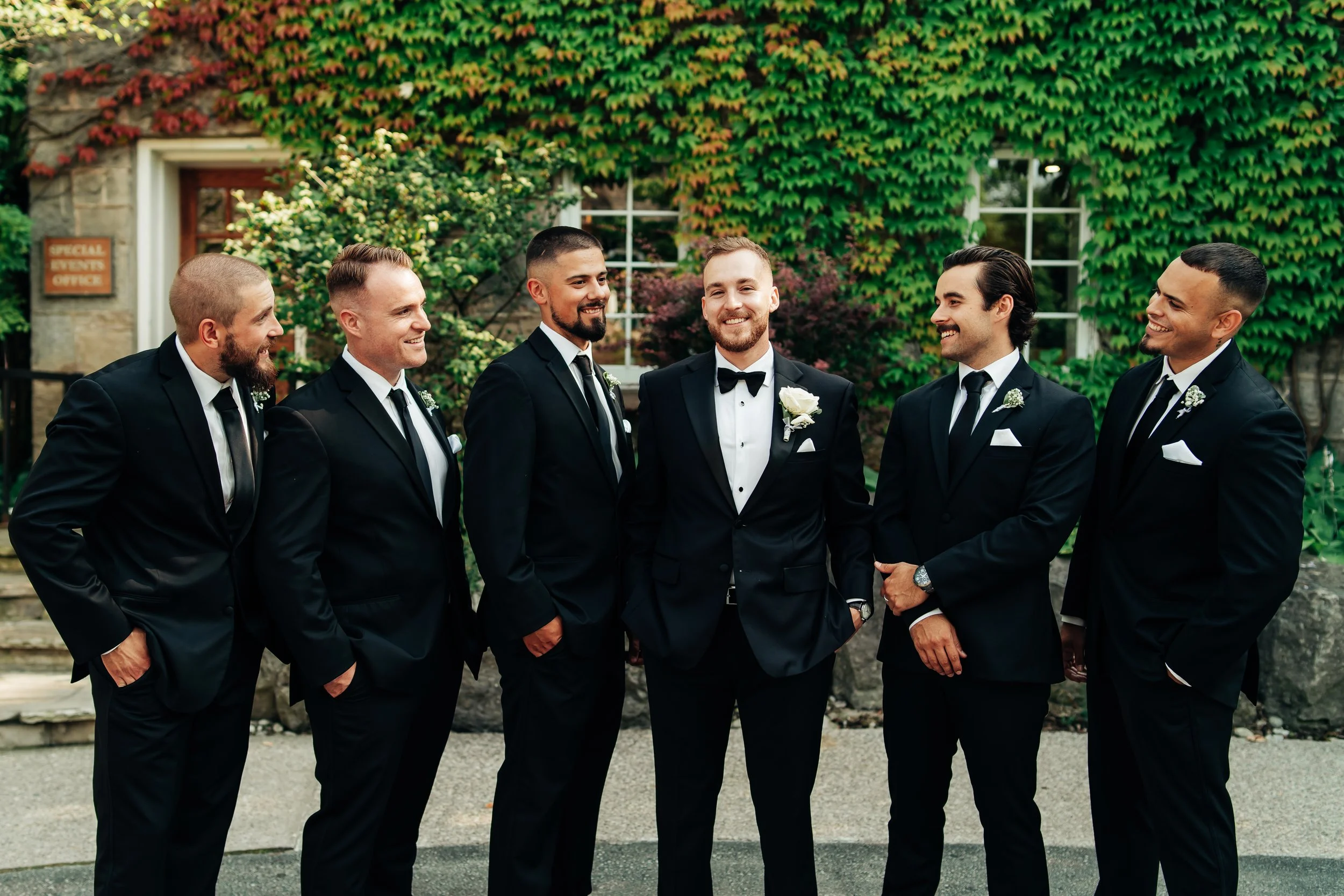 A groom in a black tuxedo with a white shirt and black bow tie, smiling, standing among six groomsmen in black suits, white shirts, and black ties, outdoors in front of a vine-covered building.