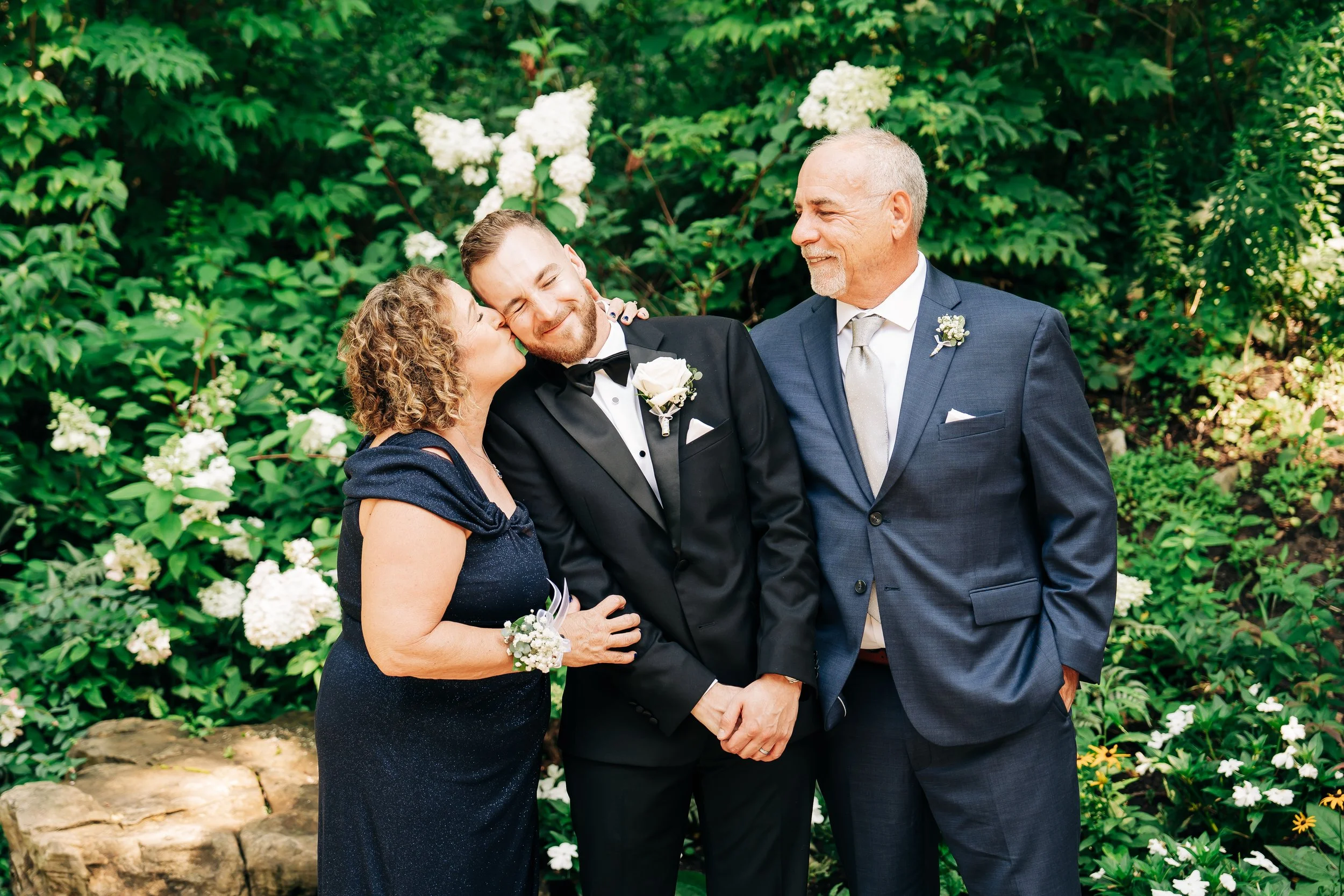 A woman in a navy dress kisses a man in a black tuxedo on the cheek, while an older man in a blue suit watches with a smile in a garden with white flowers.