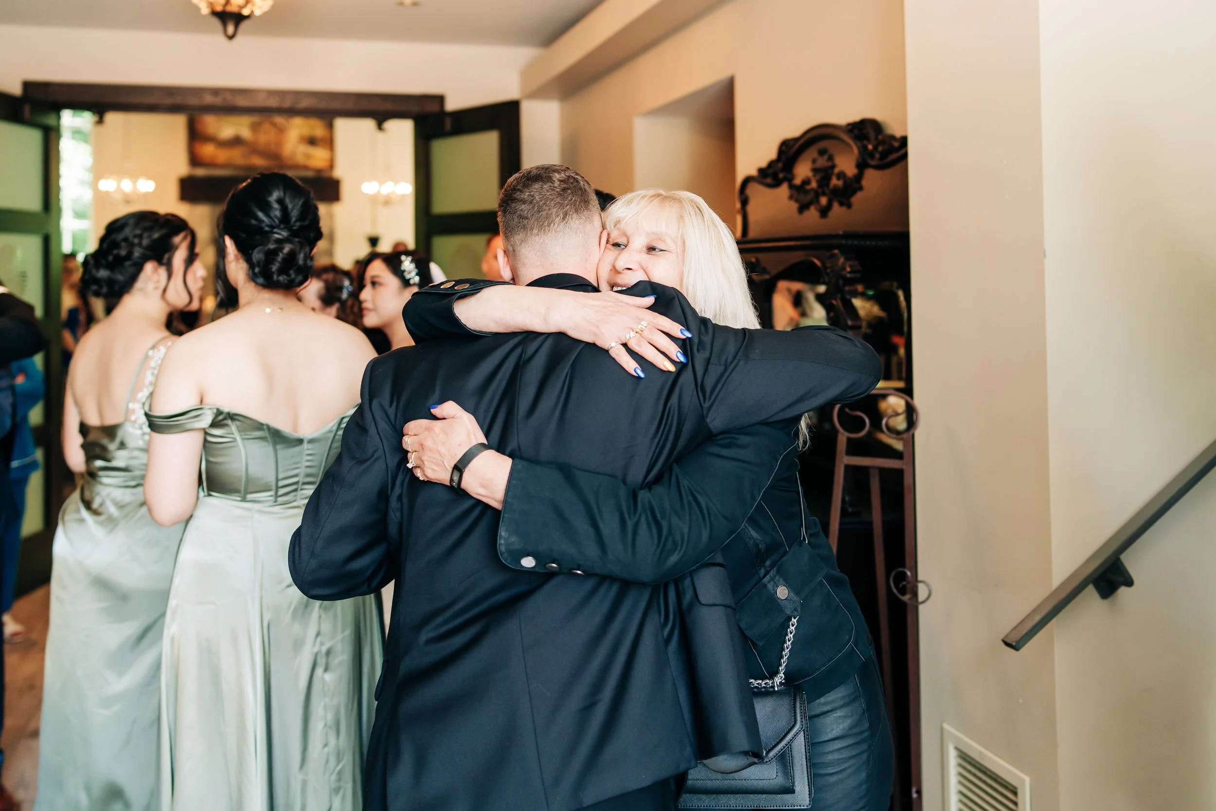 A group of women in elegant dresses in the background, with two people hugging warmly in the foreground at a celebratory event.