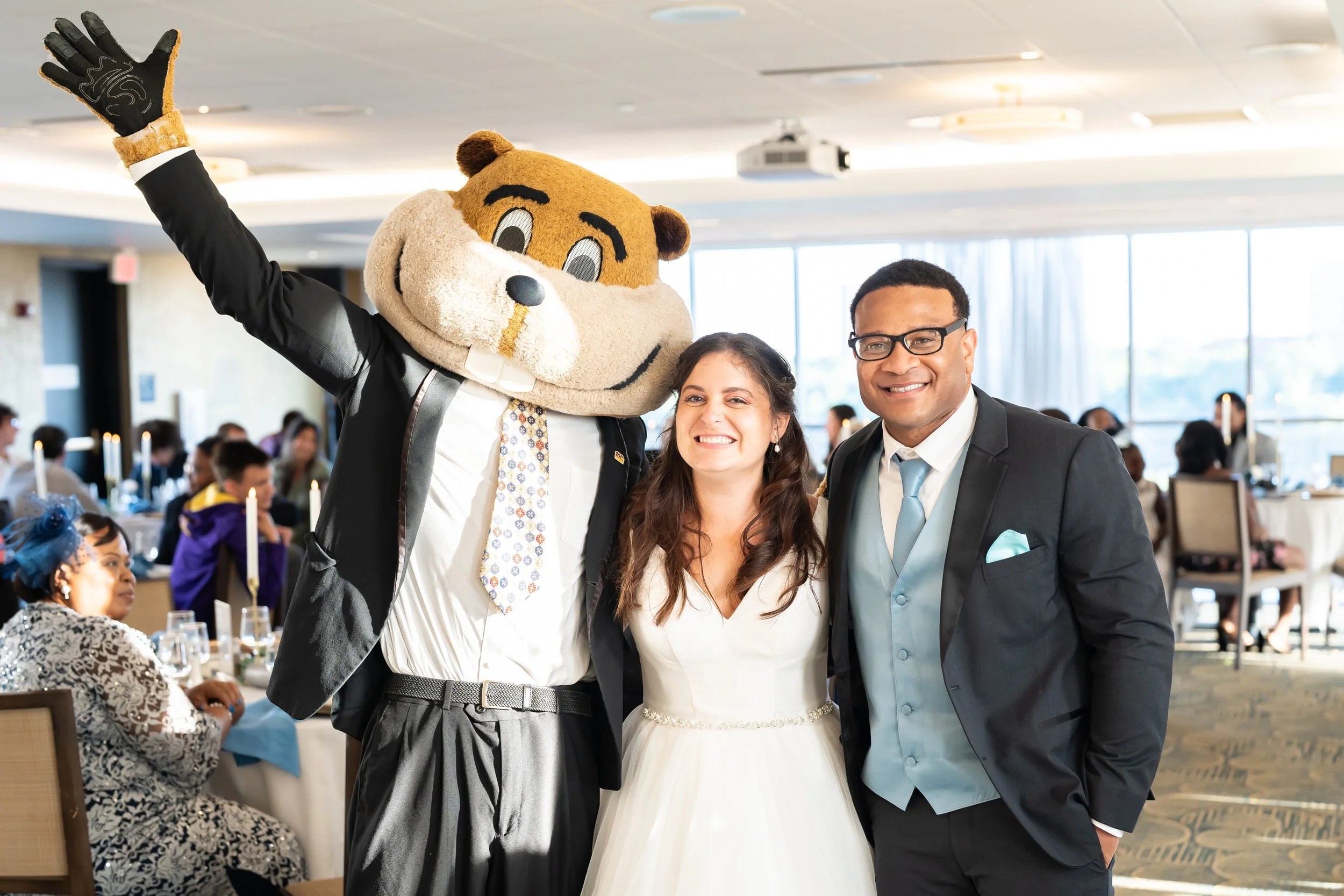 Three people with a mascot at a formal event: a person in a chipmunk costume, a woman in a wedding dress, and a man in a suit, smiling together.