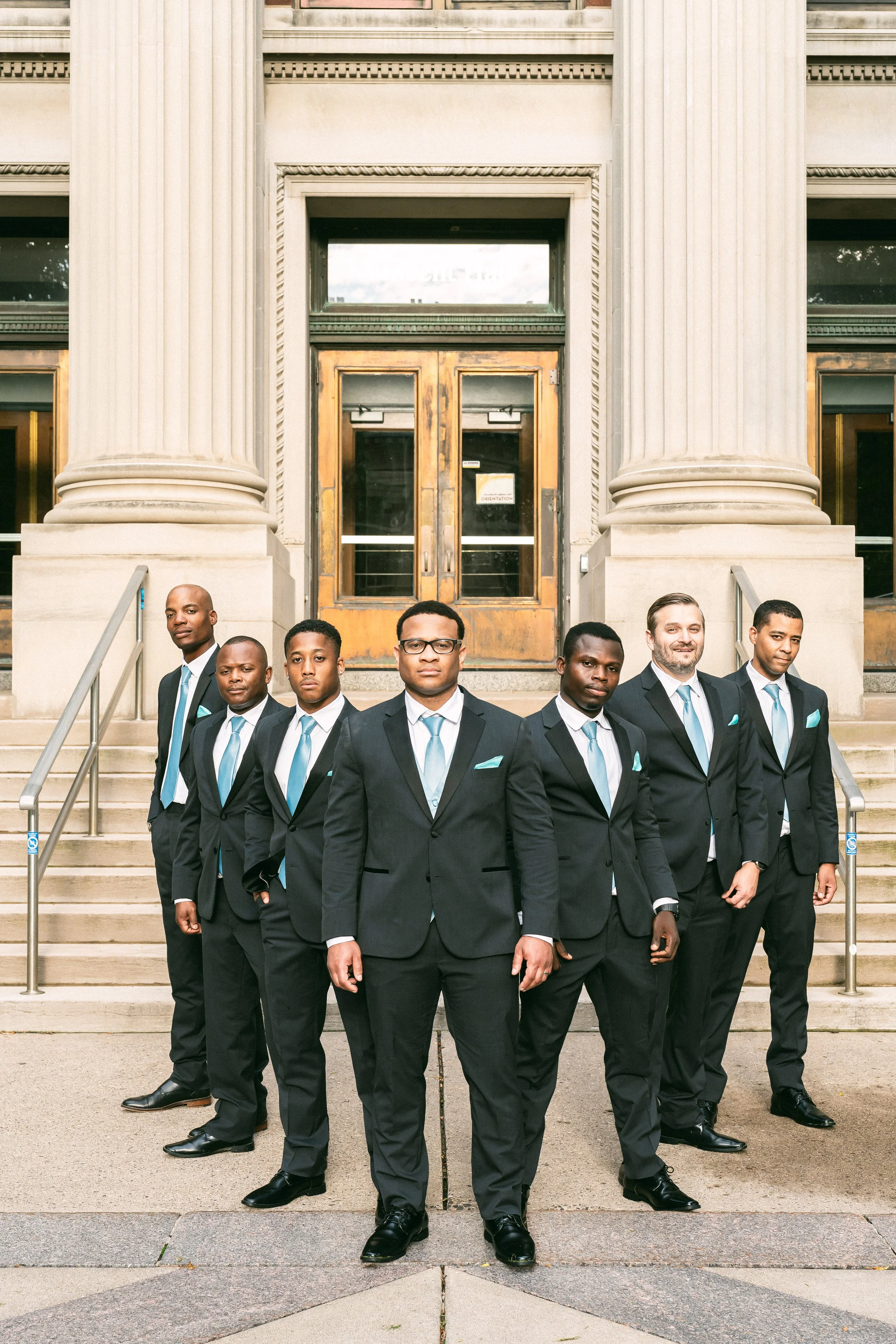 Group of eight men dressed in dark suits with light blue ties, standing on steps in front of a building with large columns and wooden double doors.