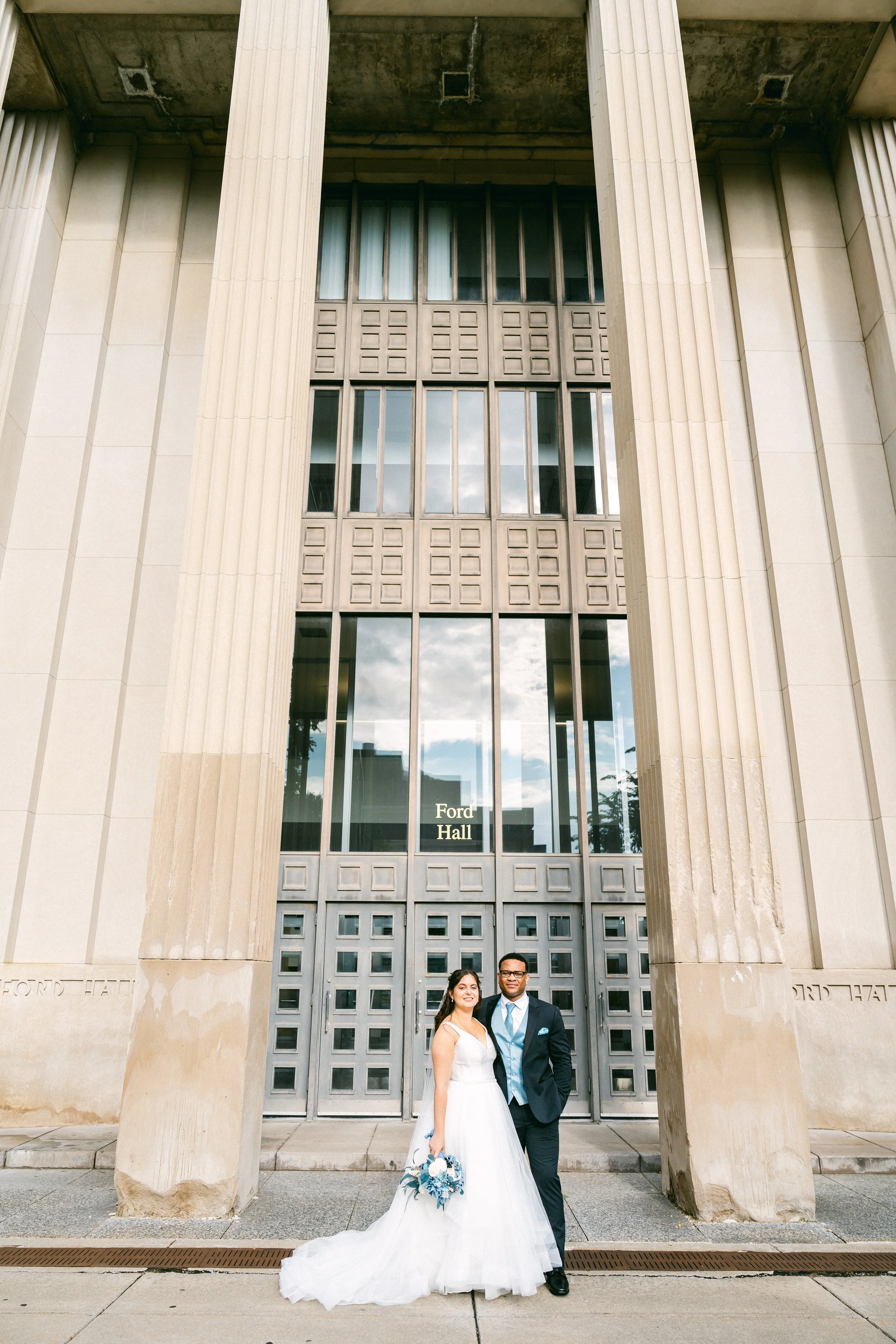A newly married couple in wedding attire standing in front of Ford Hall building. The bride is holding a bouquet, and the groom is dressed in a suit and tie. They are smiling and posing for the photo.