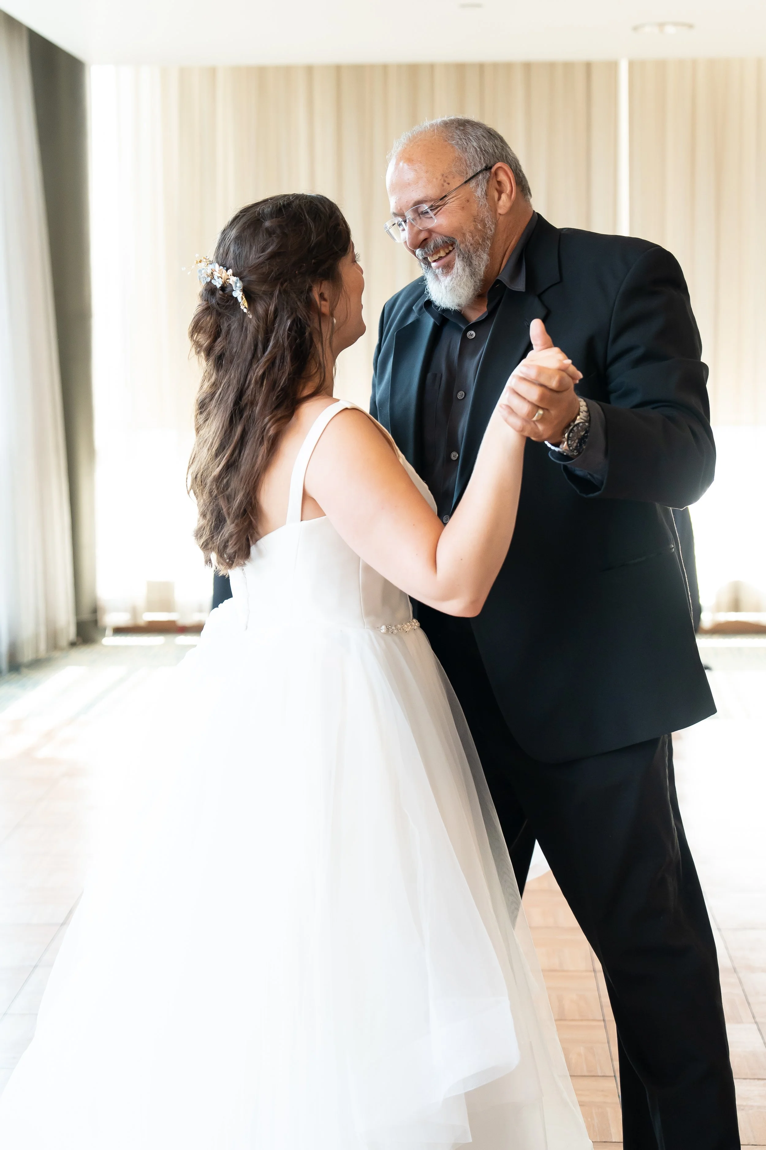 A bride and an older man dancing together in a bright room with cream curtains.