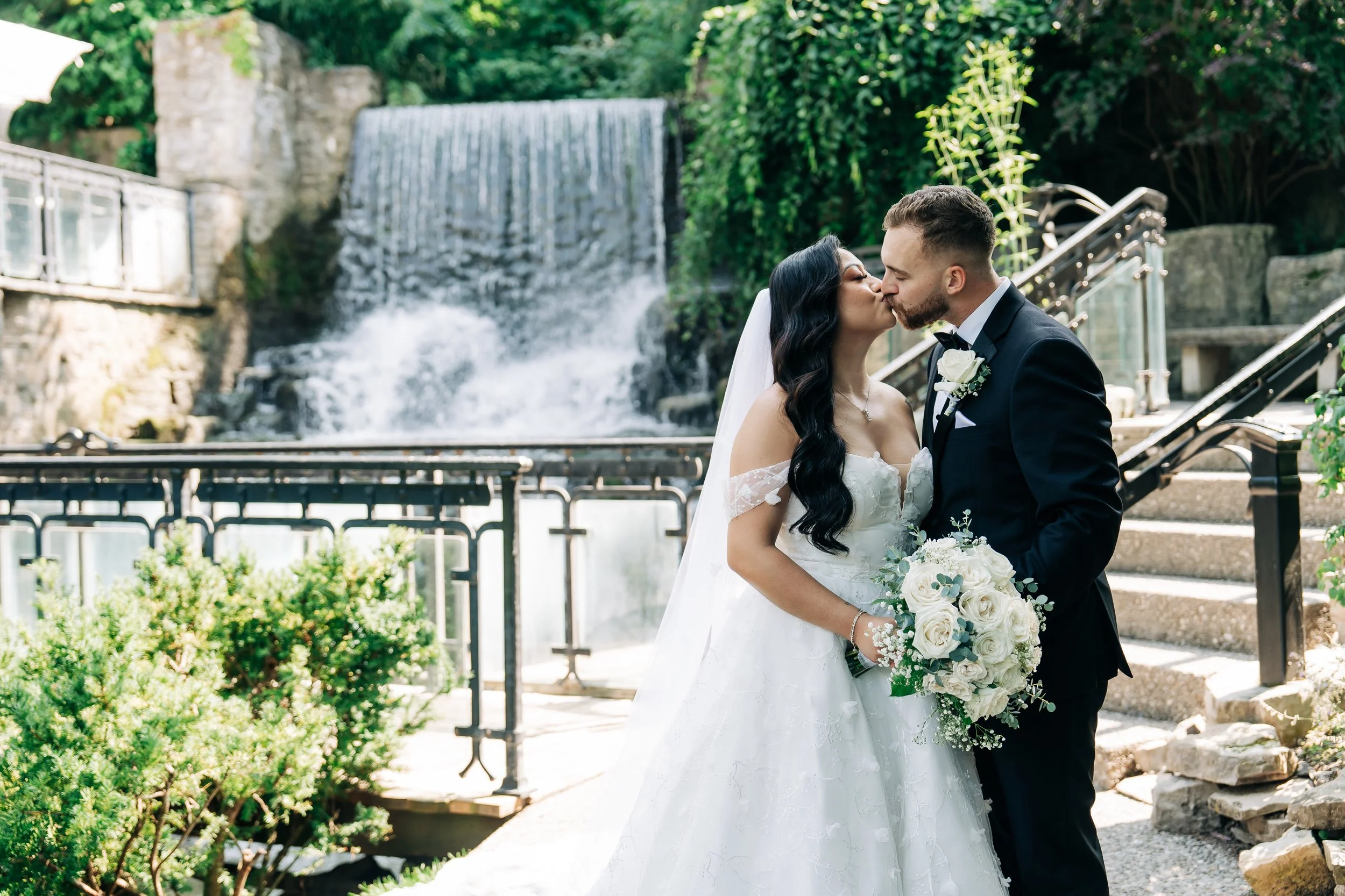 A bride and groom share a kiss outdoors near a waterfall and bridge, with greenery and stone steps around them.