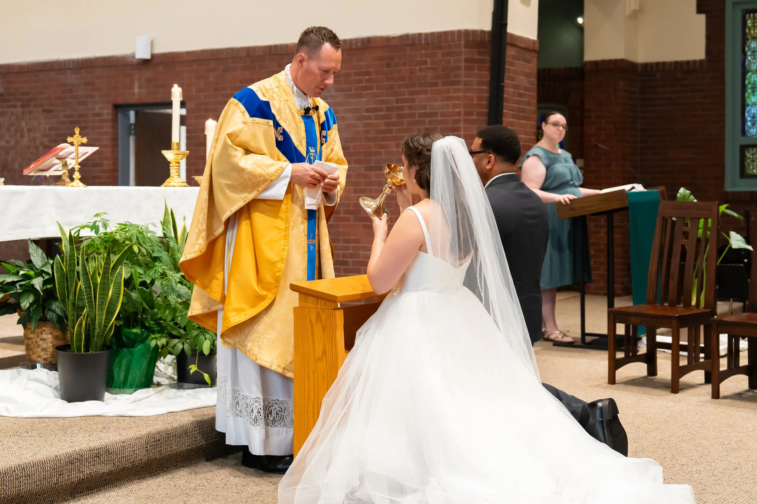 A bride and groom are kneeling in front of a priest during a wedding ceremony in a church. The priest is wearing a gold and blue vestment, and the bride is holding a chalice. In the background, a woman is standing at a lectern, and two green potted p