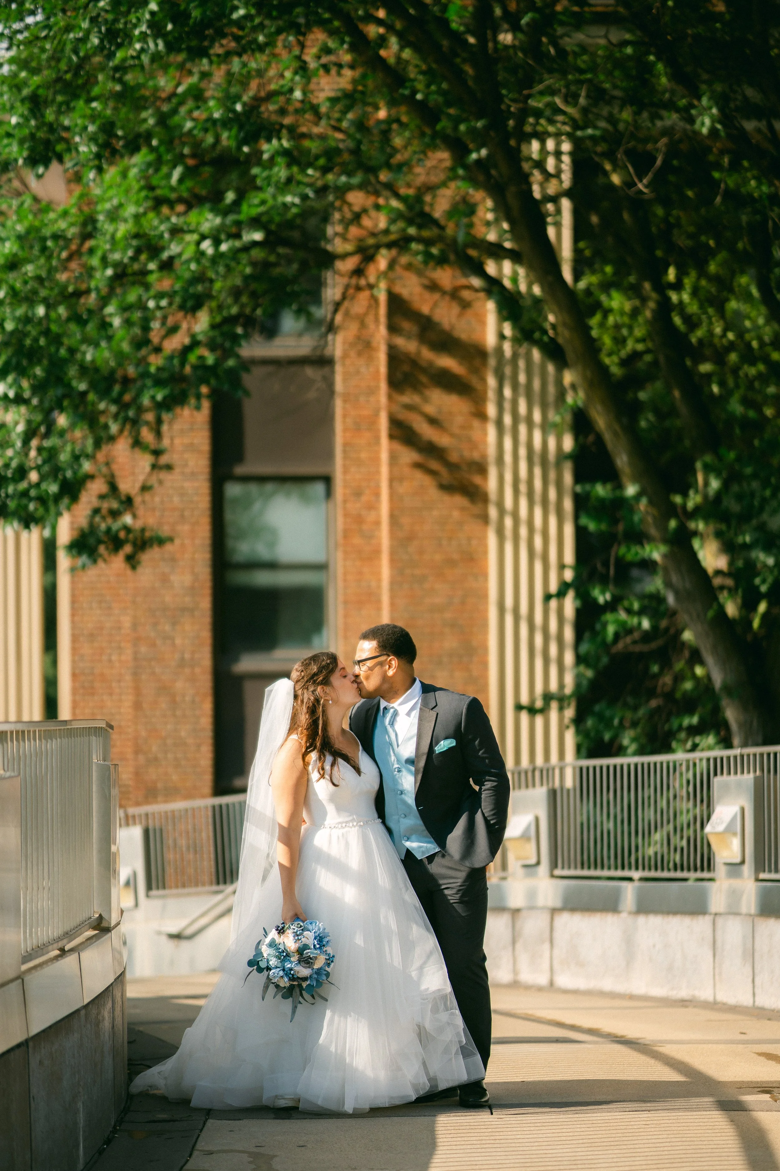 A newlywed couple sharing a kiss outdoors on a sunny day, with the bride holding a bouquet of blue and white flowers and wearing a white wedding gown with a veil, and the groom in a dark suit with a light blue vest and tie, on a city walkway with tre
