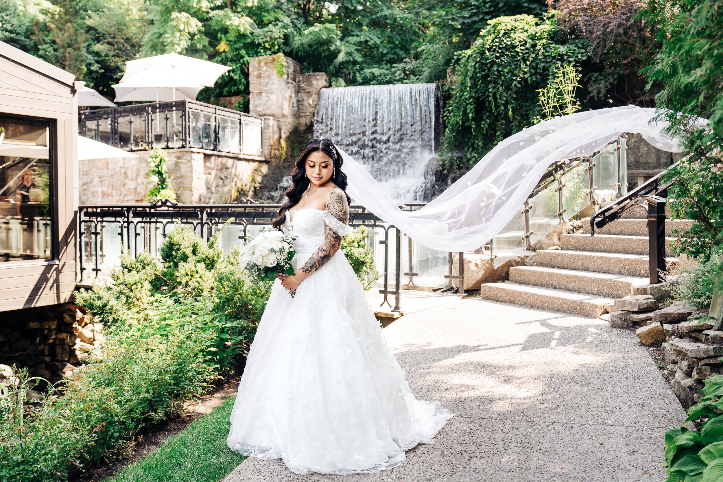 Bride in a white wedding dress holding a bouquet, standing outdoors near a waterfall with lush greenery and a staircase in the background.