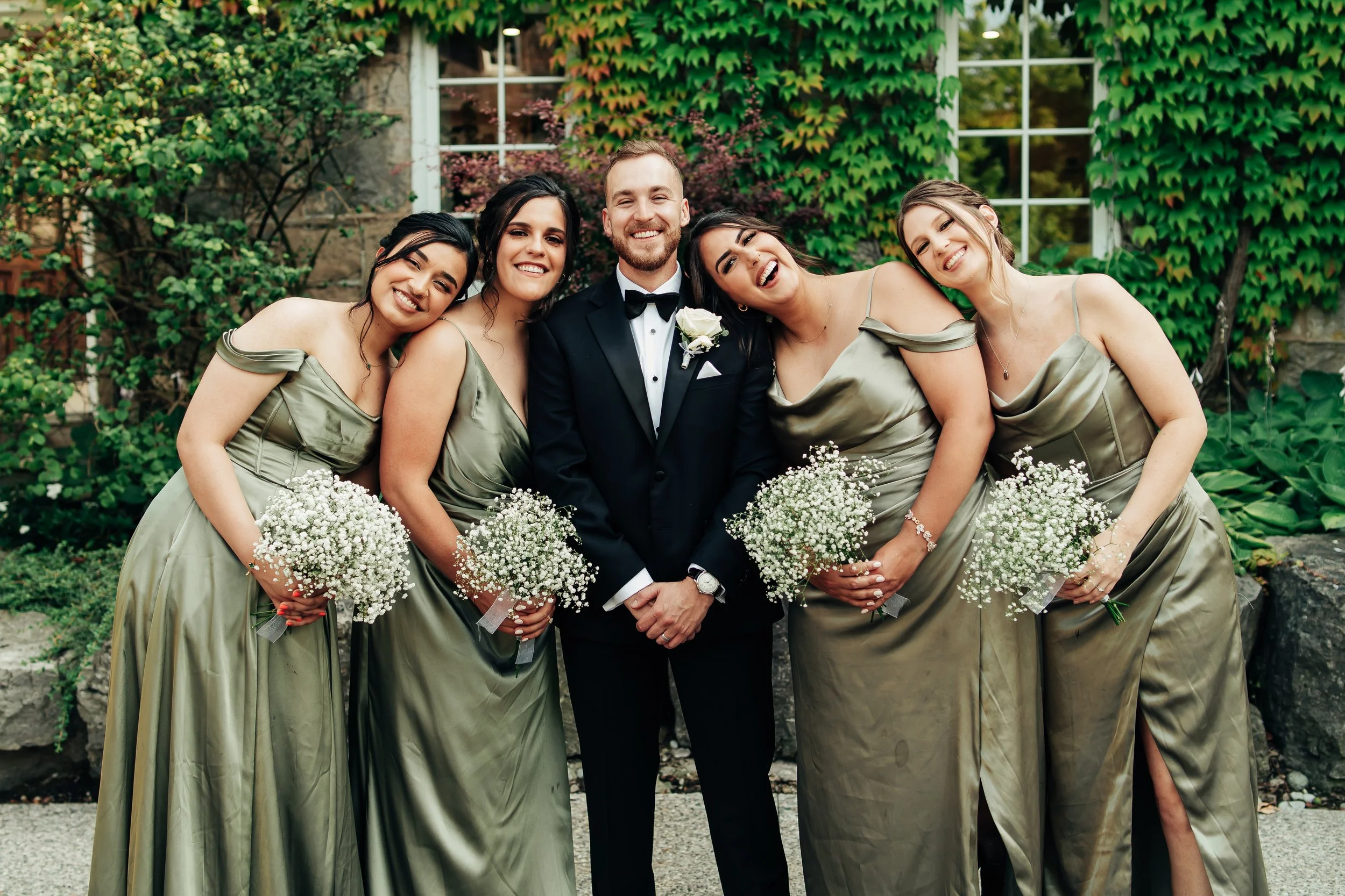 Group of five people, a groom in a tuxedo and four bridesmaids in matching satin dresses, smiling and holding bouquets of white baby's breath flowers, outdoors in front of a lush green ivy-covered wall.