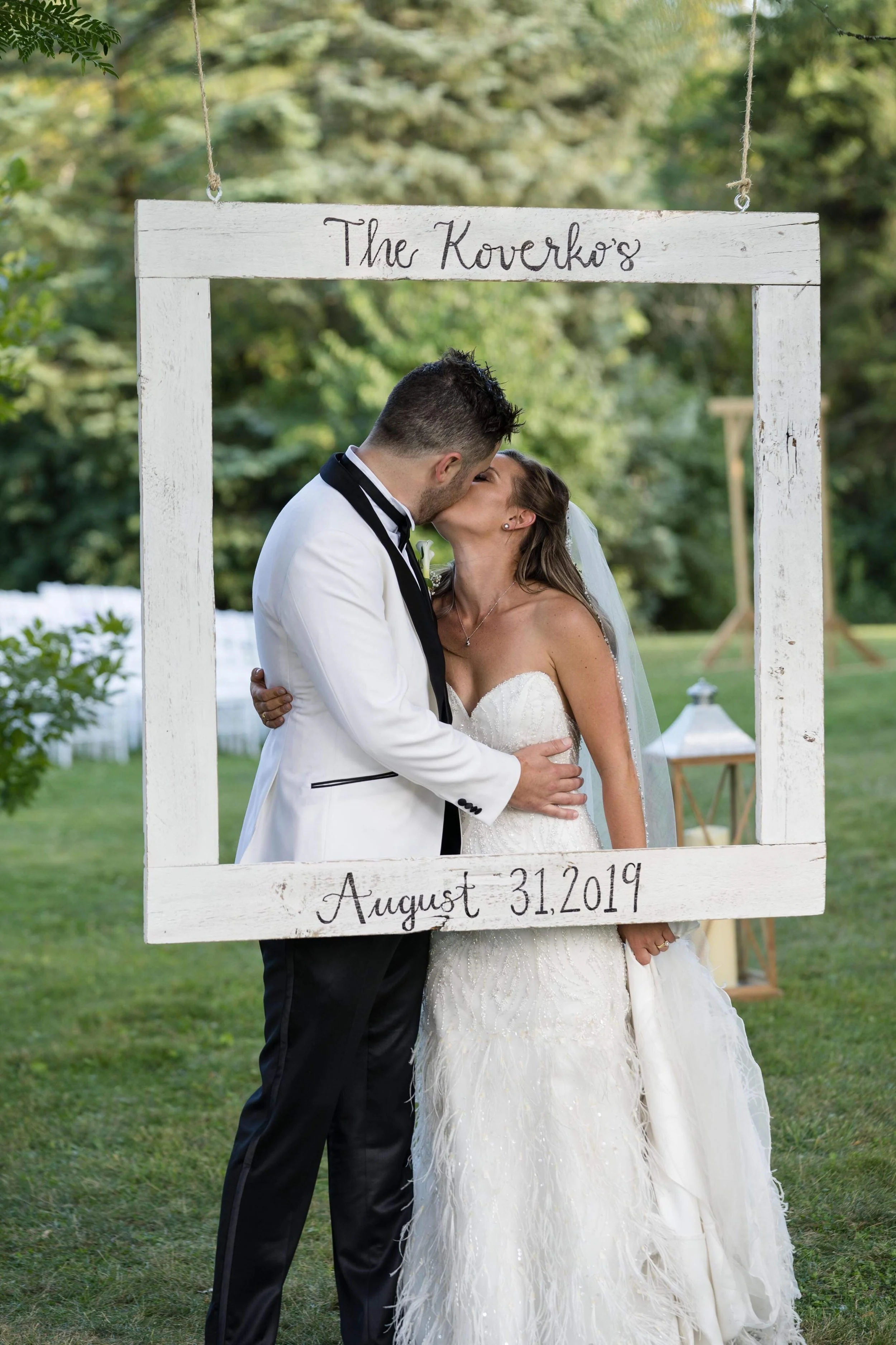 A bride and groom sharing a kiss during their outdoor wedding, framed by a white wooden square with handwritten text that reads 'The Kovarko's' at the top and 'August 31, 2019' at the bottom. Green trees and grass are in the background.