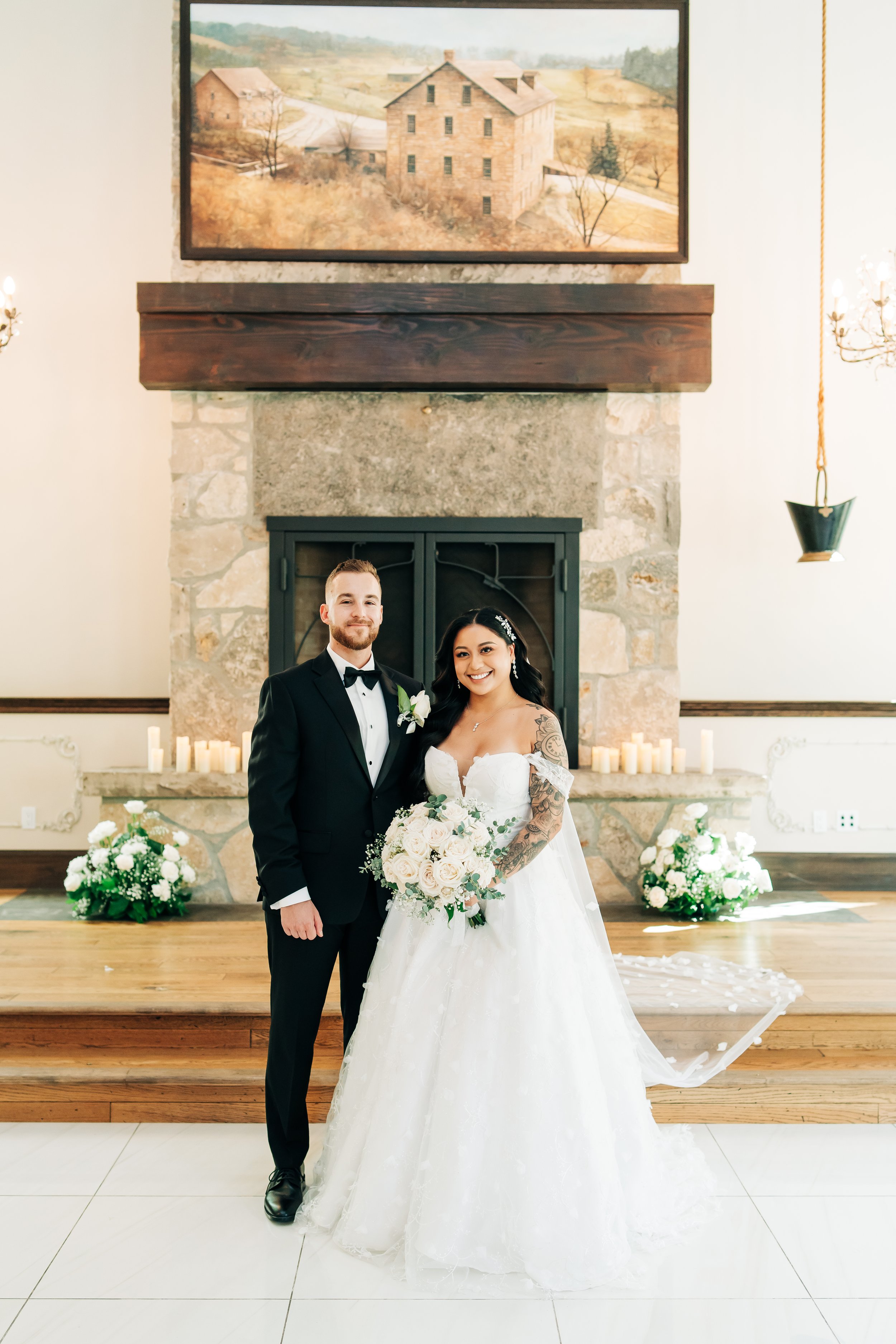A bride and groom posing indoors in front of a stone fireplace with a large painting of a rustic house hanging above it. The bride is holding a bouquet of white roses and greenery, wearing a white wedding gown with tattoos visible on her right arm. T