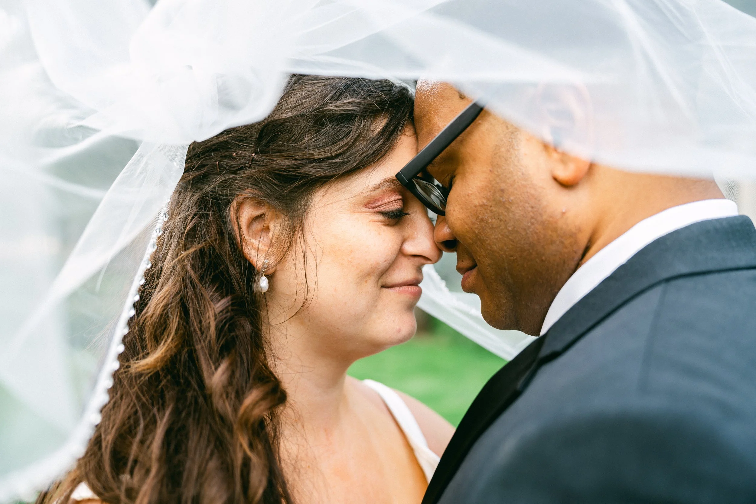 A bride and groom with foreheads touching under a white veil, smiling with eyes closed during a wedding.