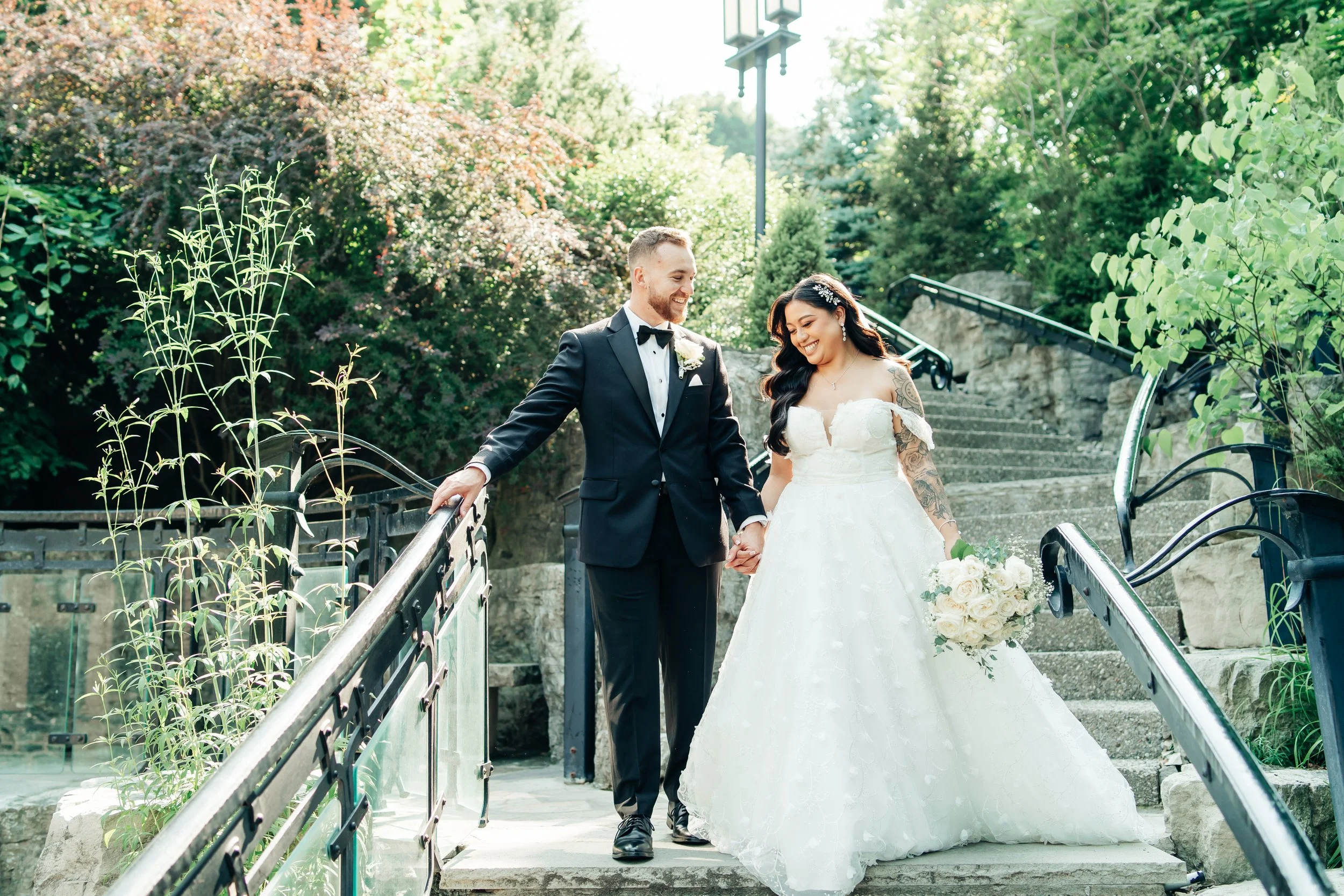 A wedding couple, a groom in a black tuxedo and a bride in a white wedding dress holding hands on outdoor stairs surrounded by greenery.