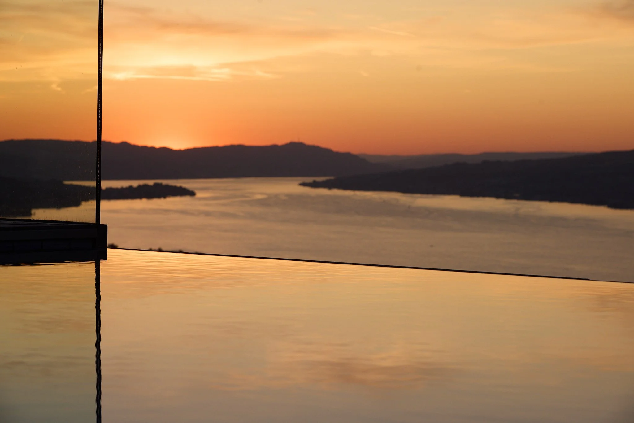 Sunset over a river with mountains in the background, seen from a reflective surface, possibly a pool or glass barrier.