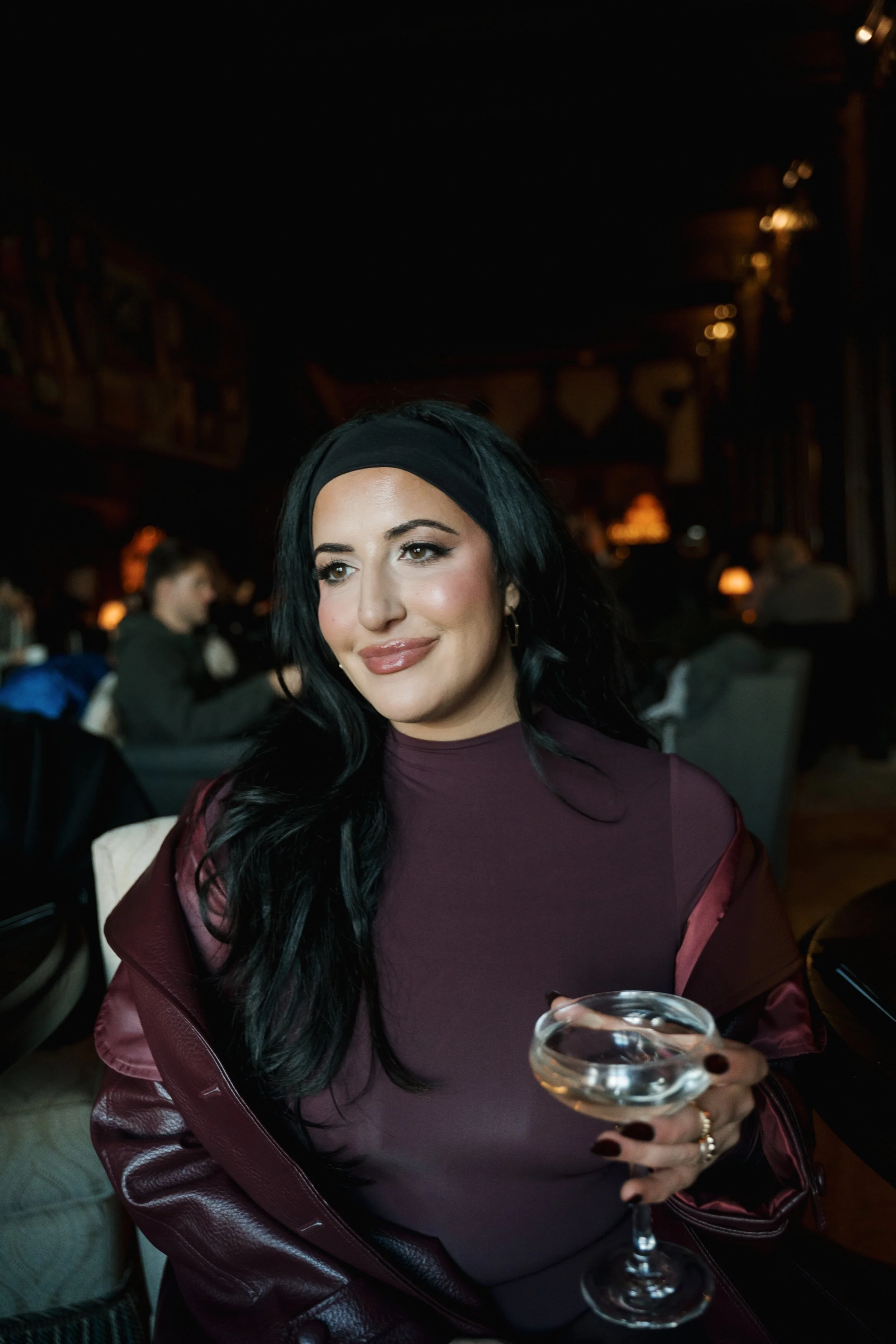 A woman with black hair, wearing a black headband and maroon lipstick, sitting in a dimly lit restaurant or bar, holding a cocktail glass with a clear drink and garnished with a cherry, surrounded by other patrons.