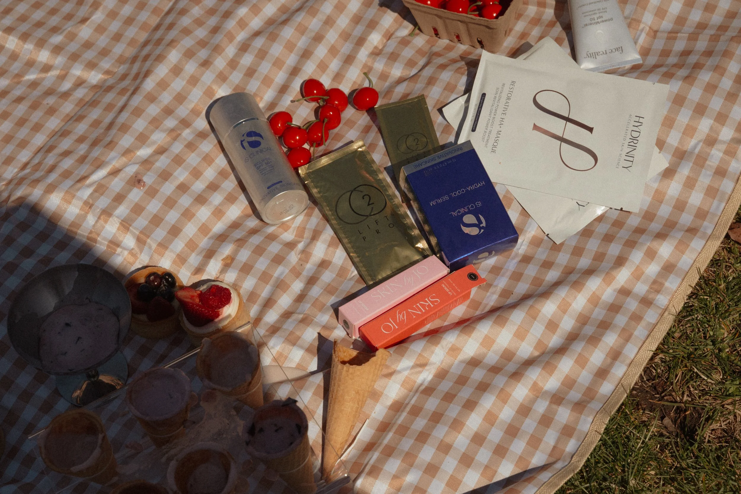 A checkered picnic blanket with various items including a bunch of cherries, a tube of skincare cream, a box of chocolate, a small cake, and skincare product boxes.