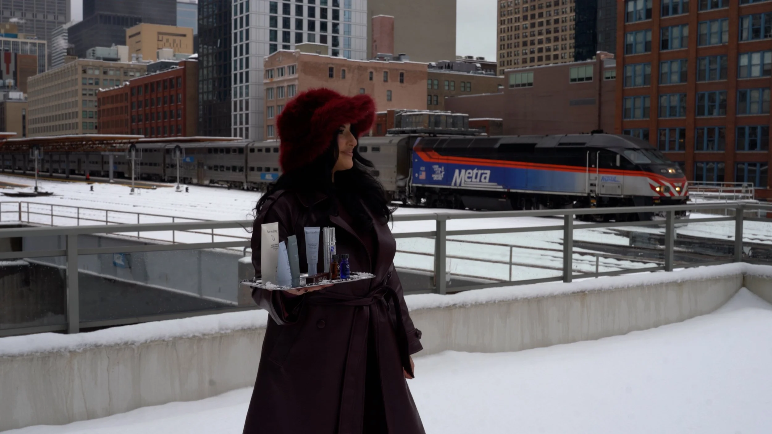 A woman in a burgundy coat and matching furry hat stands outdoors in a snowy cityscape, holding a tray of skincare products, with a Metra train and tall buildings in the background.