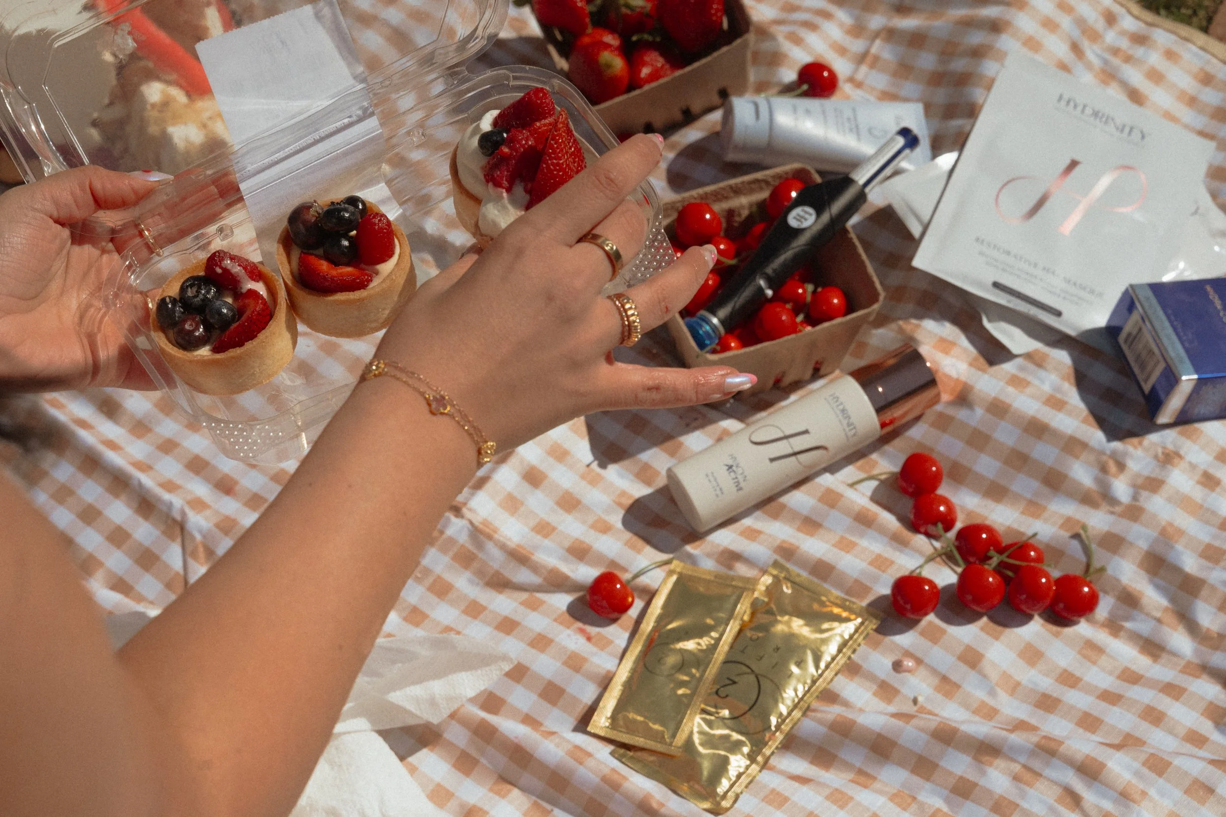 Close-up of a hand holding a small tart filled with whipped cream and topped with strawberries, blueberries, and raspberries on a checkered tablecloth surrounded by cherry tomatoes, beauty products, and gift boxes.