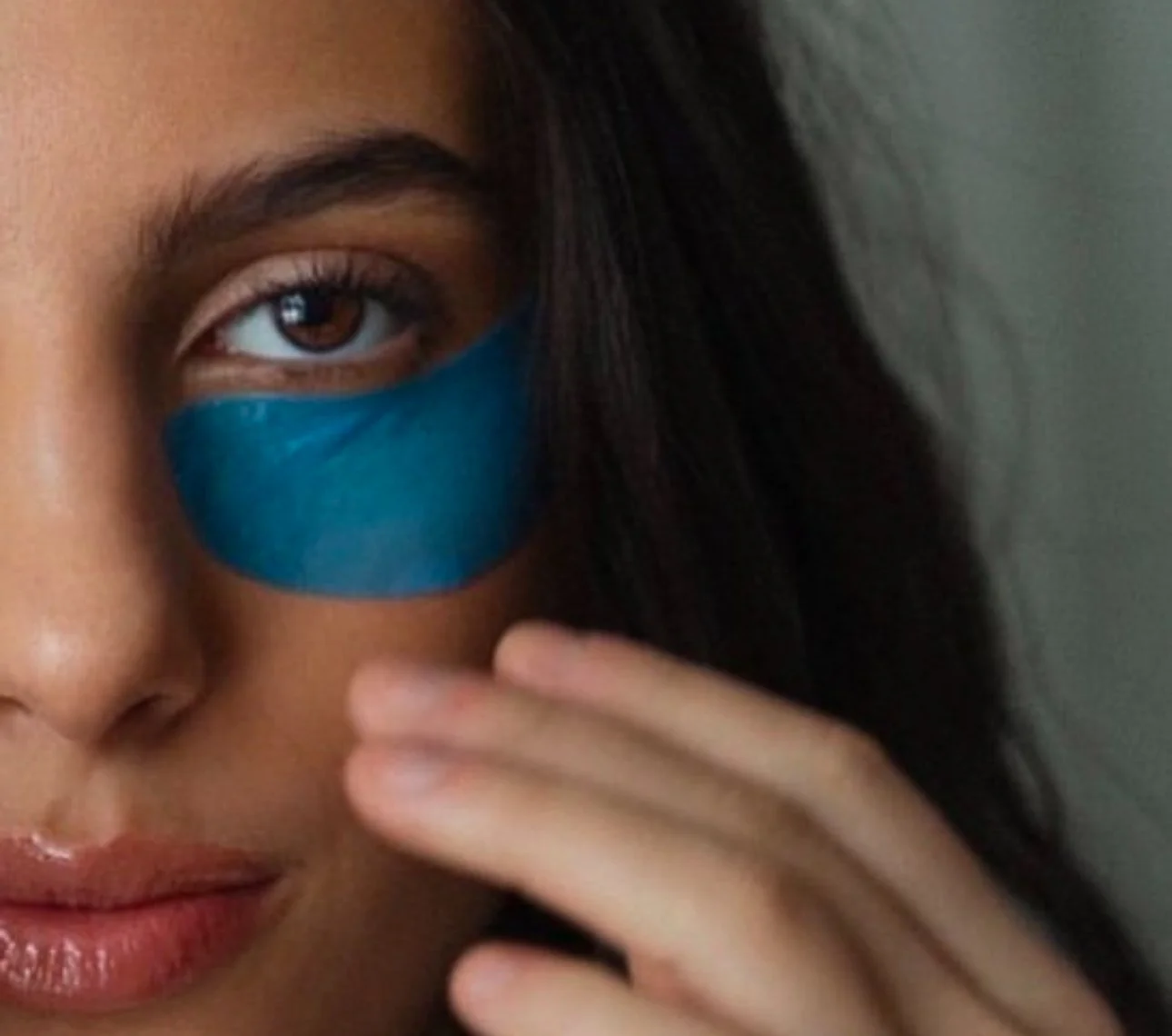 Close-up of a woman's face showing her eye and part of her nose, with a blue eye patch under her eye and her hand near her face.