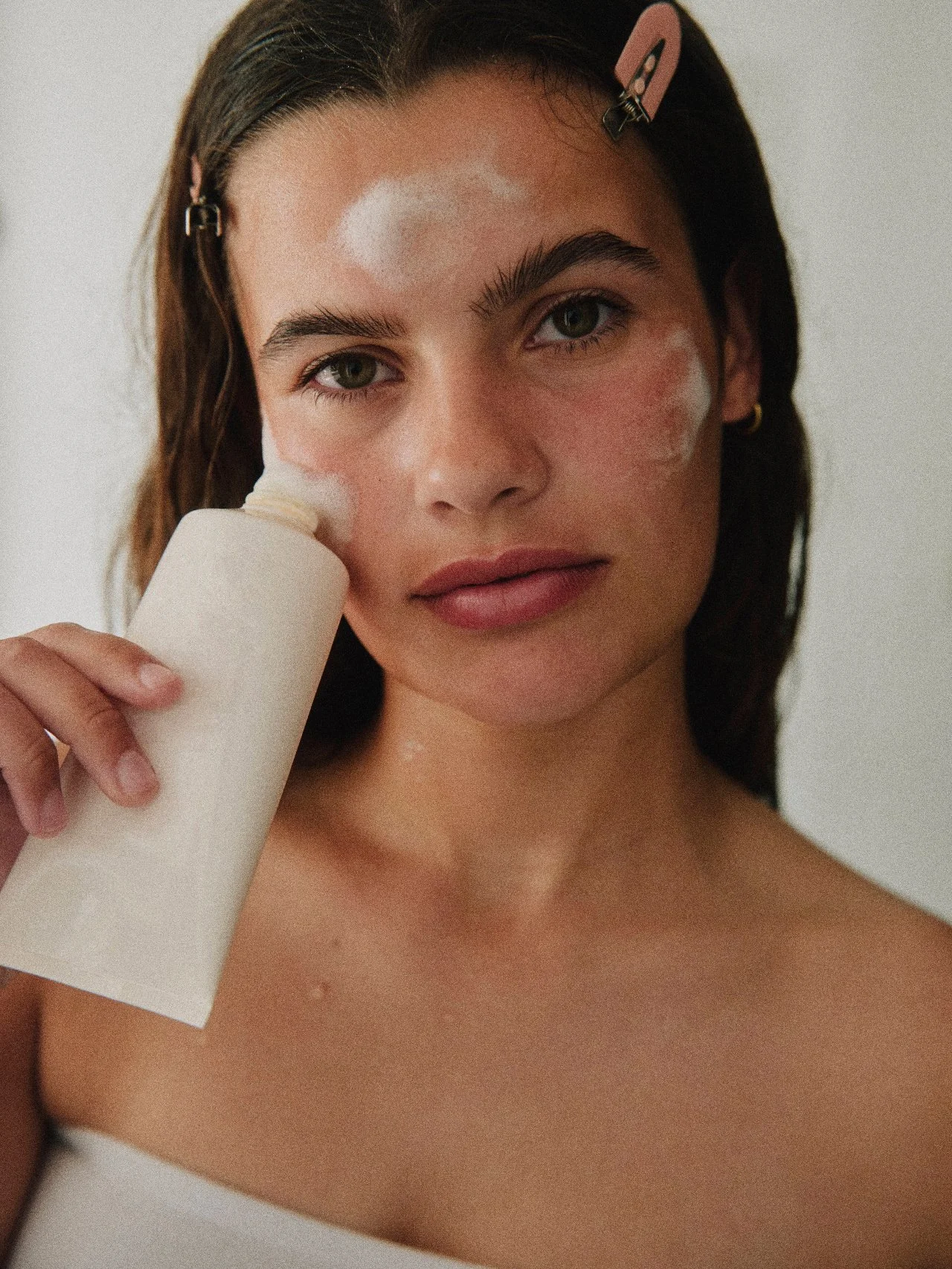 A young woman with dark hair pinned back with pink clips, holding a roll of white toilet paper to her face, with some patches of white skin ointment visible on her face.
