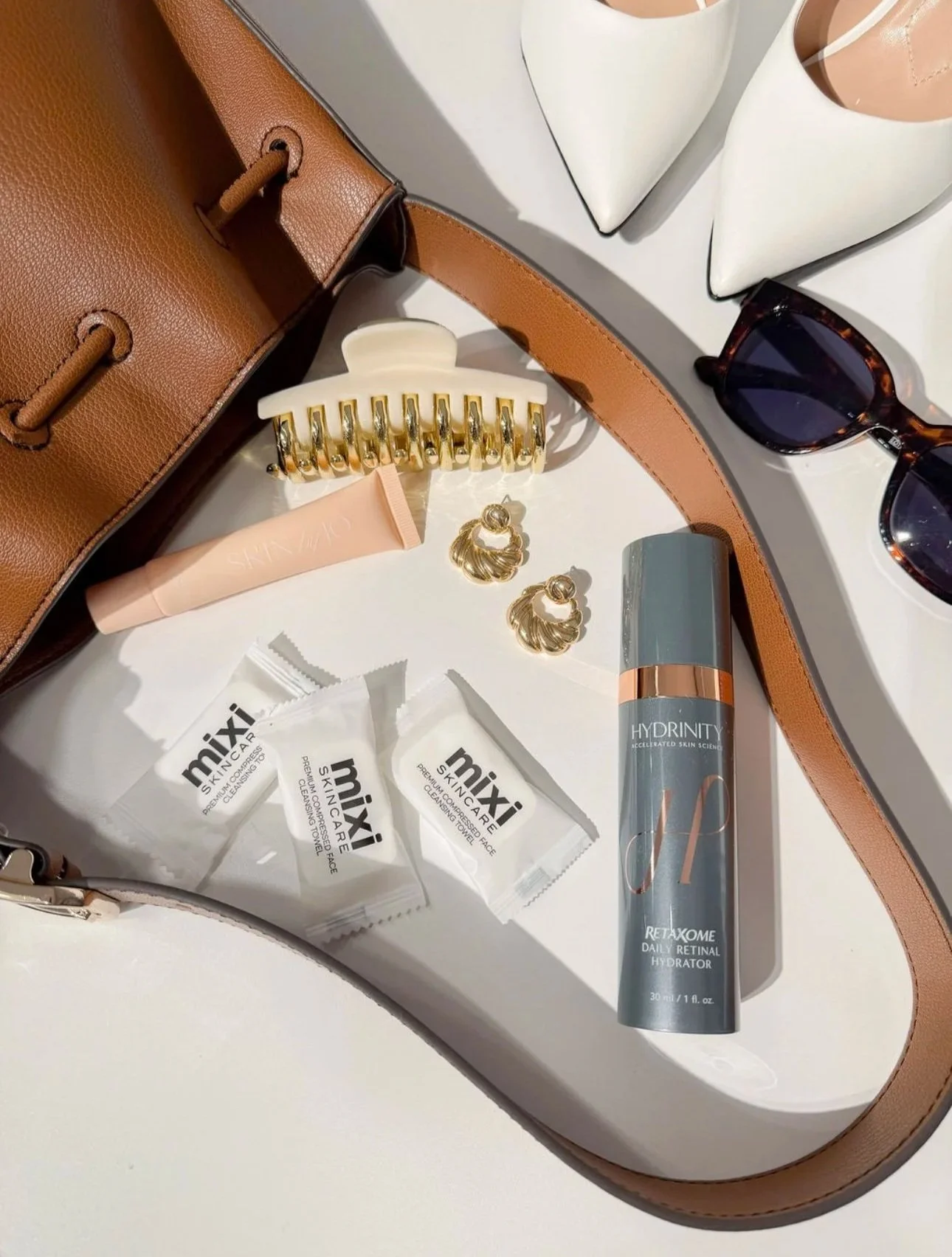 A collection of skincare and fashion accessories including a brown leather handbag, white high heels, tortoise shell sunglasses, gold hoop earrings, a cream-colored hair claw clip, a pink facial cream, a gray cosmetic spray bottle, and three packet samples labeled 'mixi SKINCARE'.