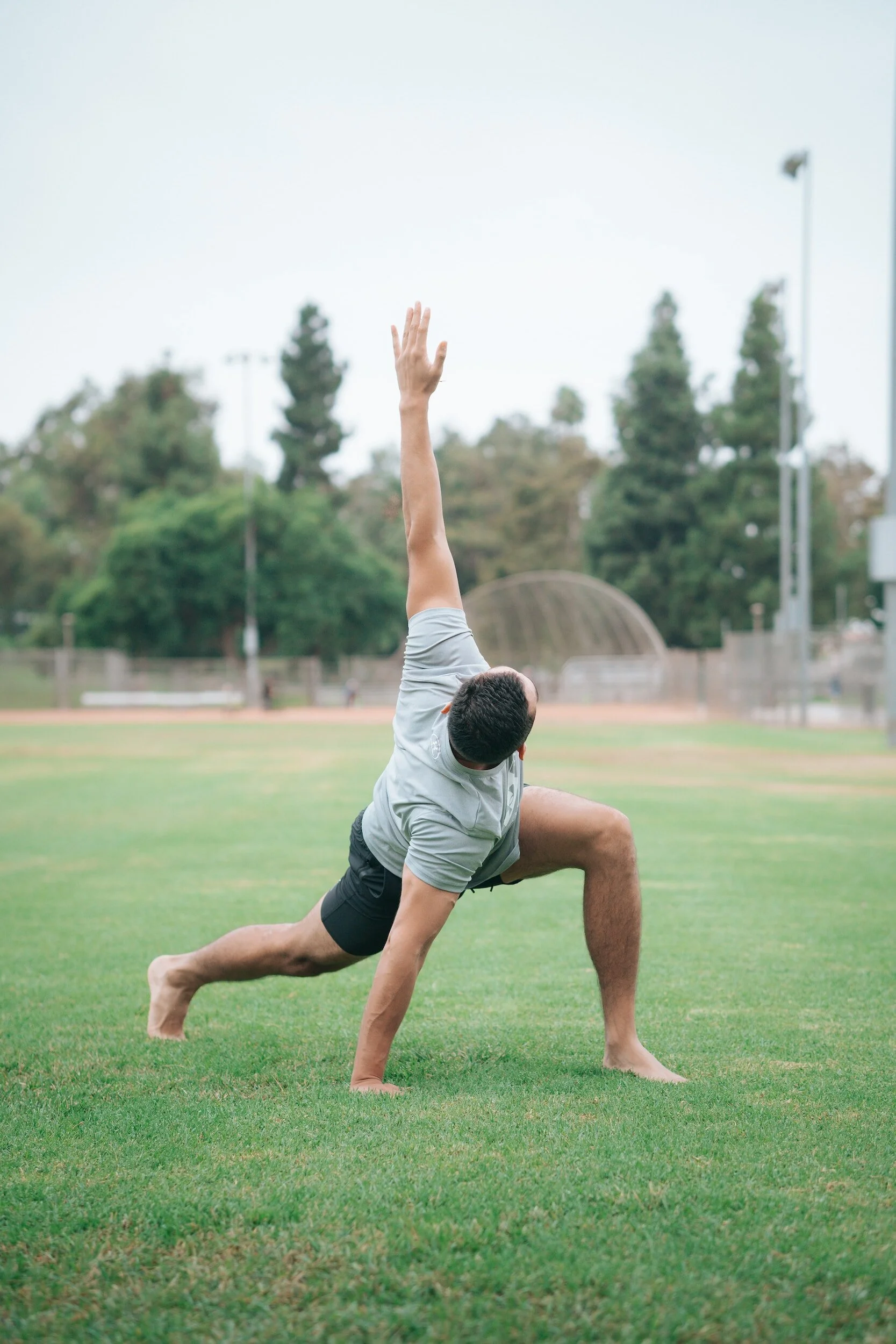 A man practicing yoga on a grassy field, in a low lunge pose with one arm reaching upward.