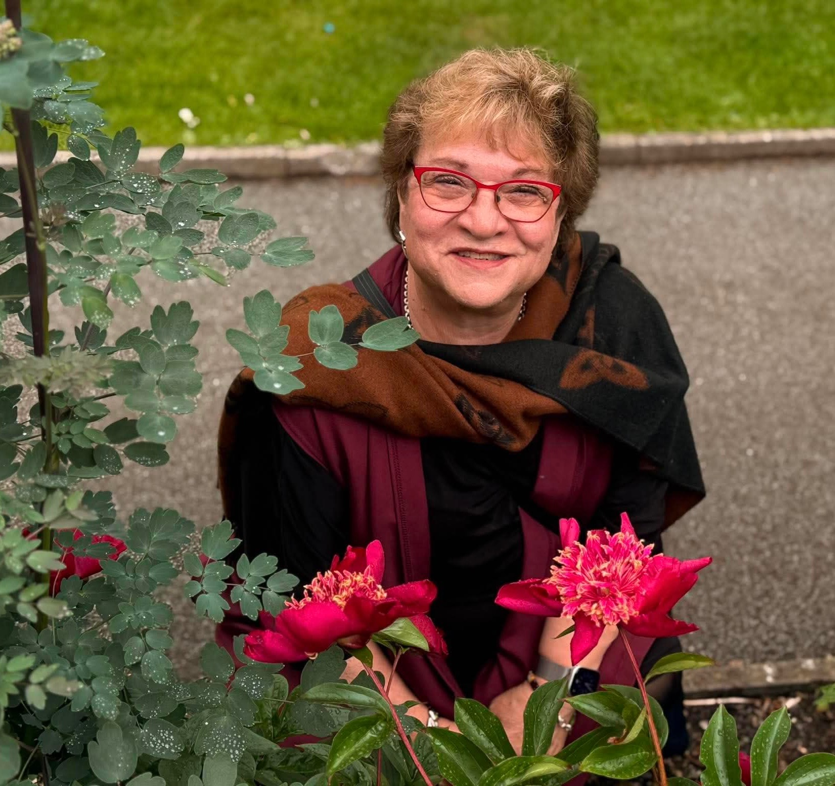 A smiling woman with short brown hair and glasses wearing a blue top and necklace, outdoors with green trees and blue sky in the background.