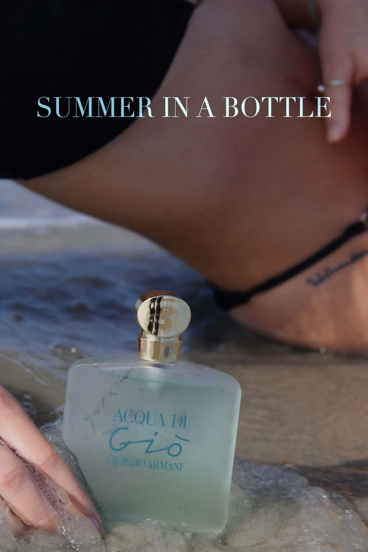 A person with a tattoo on their waist holding a bottle of Giorgio Armani Acqua di Giò perfume at the beach near the water