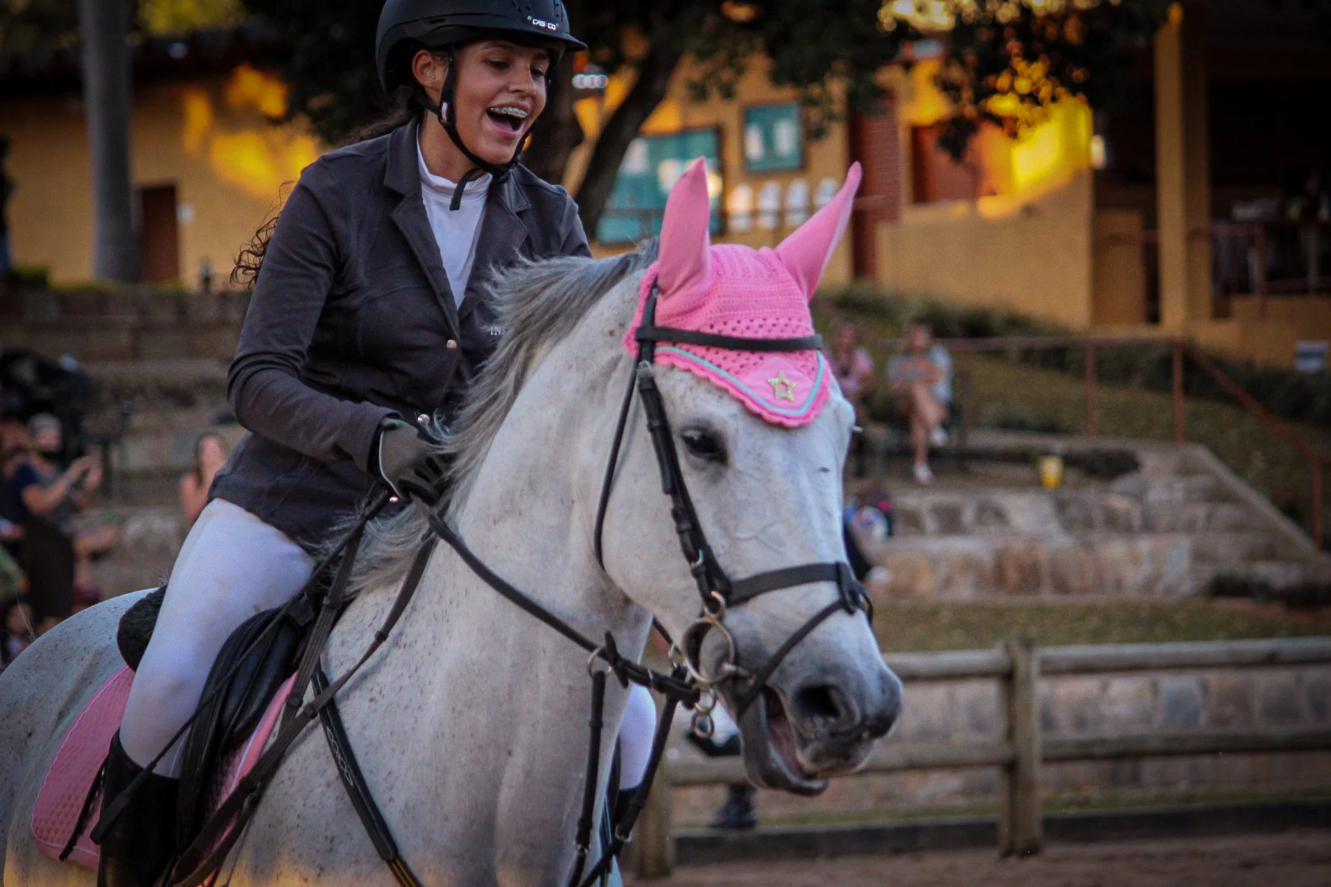 A girl riding a white horse wearing a pink knit horse hood and a black riding helmet, smiling during a sunset event at an outdoor stable.