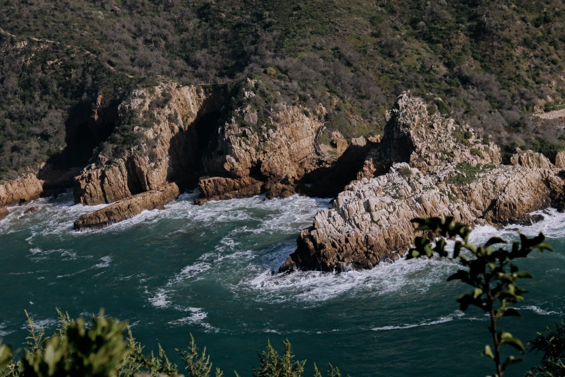 A rugged coastline with large rocks and crashing waves, backed by green hills and shrubbery.