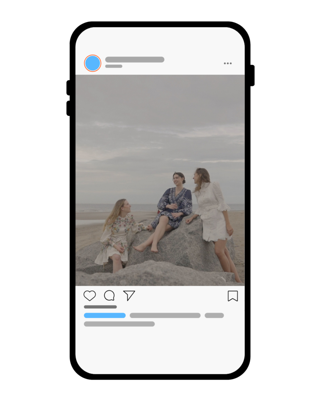 Three women on a beach, sitting on and next to large rocks, dressed in floral and white clothes, smiling and talking under a cloudy sky.