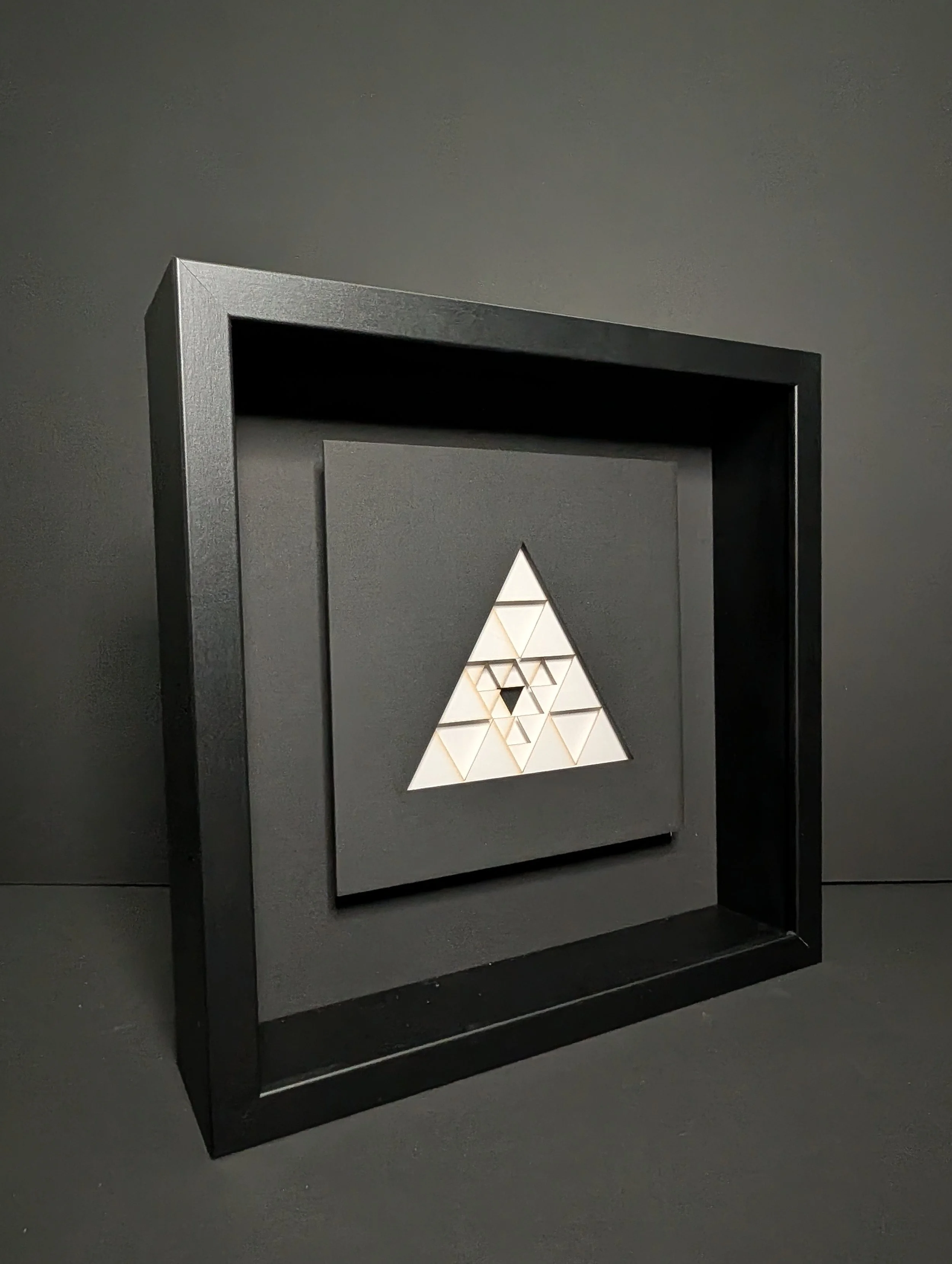 A framed geometric artwork with a large white triangle containing smaller interconnected triangles and a black inverted triangle at the center, on a dark background.