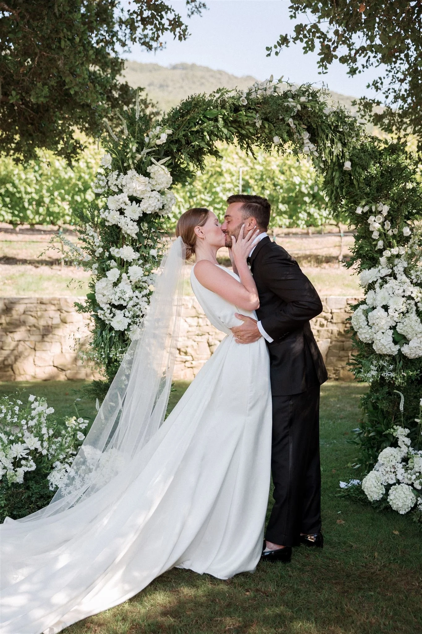 A bride and groom kiss under a floral arch outdoors on a sunny day.