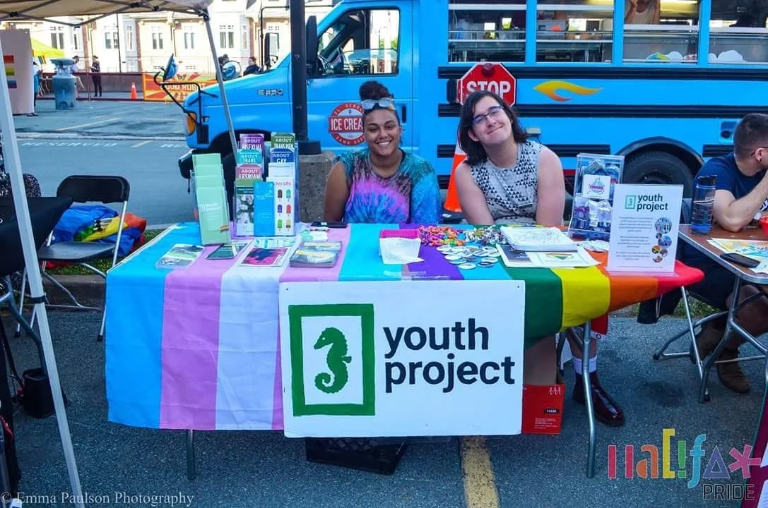 There are two people sitting at an information display table during the Halifax Pride Festival. The table is covered with the rainbow and trans flags.