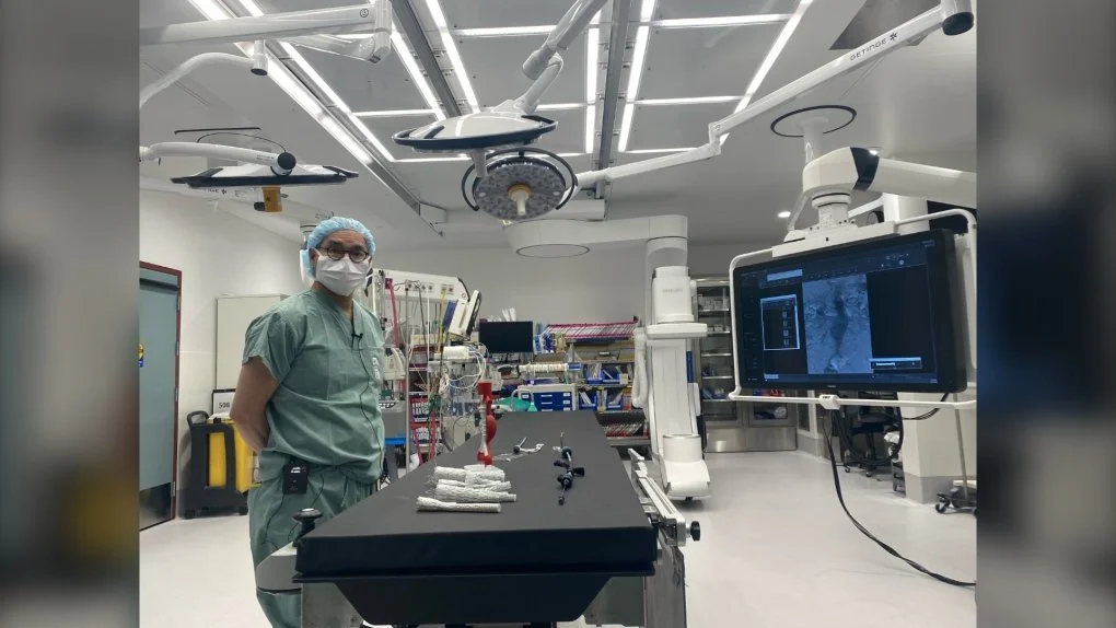 This photo shows the inside of the operating room.  There is someone in scrubs standing by a table with surgical items on it, with lots of lights above and a screen beside it.