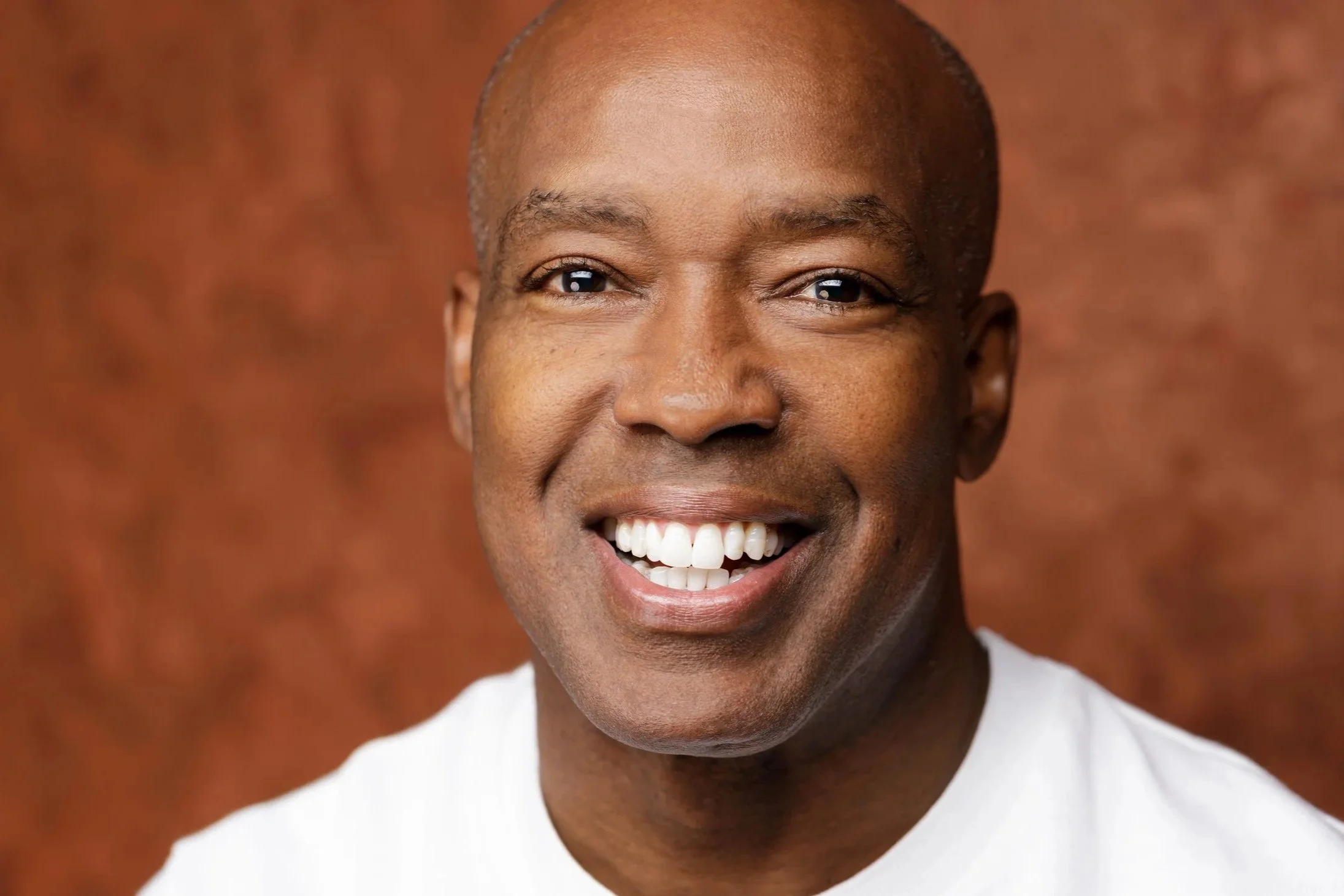 A smiling African American man with a shaved head, wearing a white shirt, against a brown textured background.