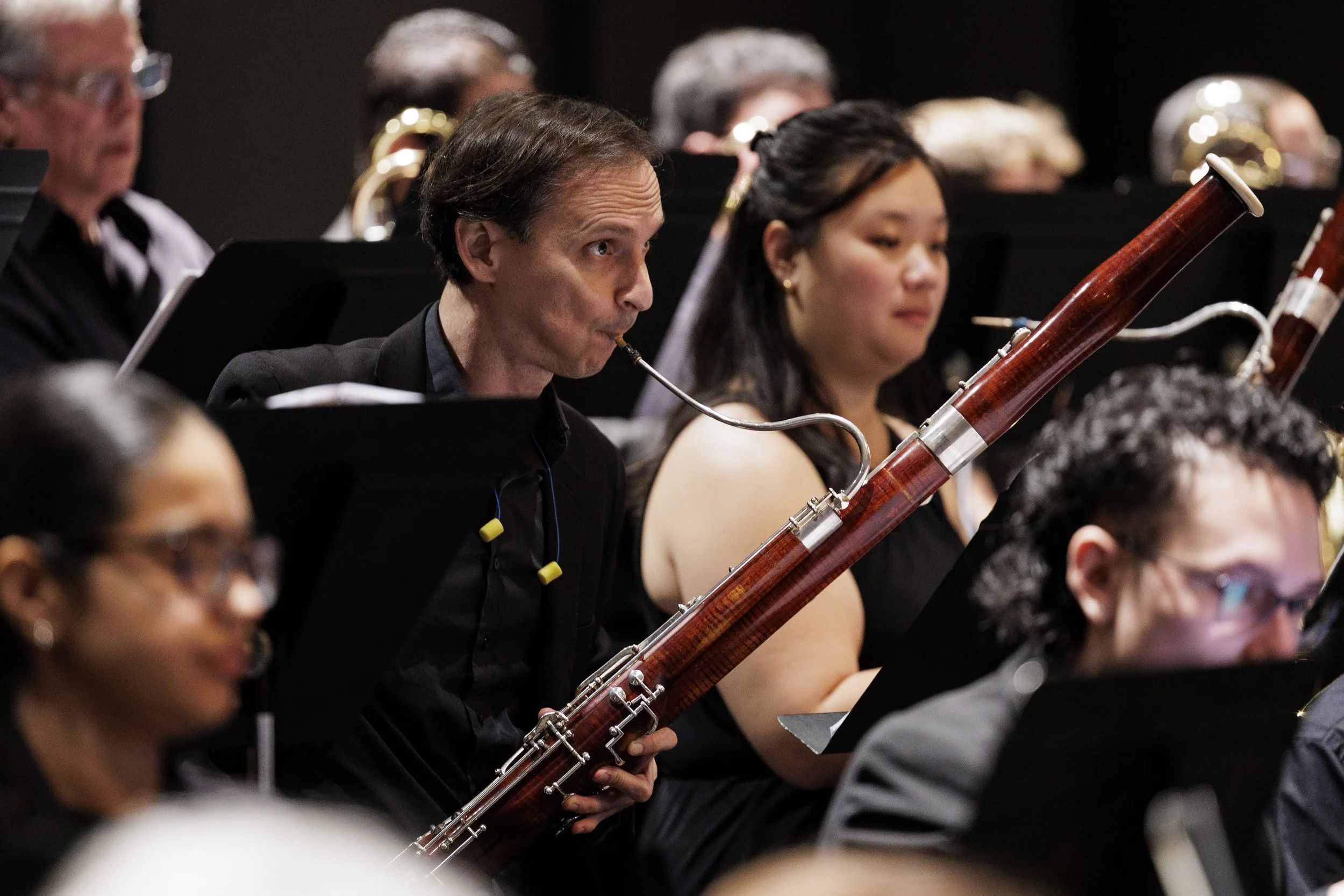 Orchestra musicians performing, focusing on a man playing a bassoon, with others playing woodwind instruments around him.