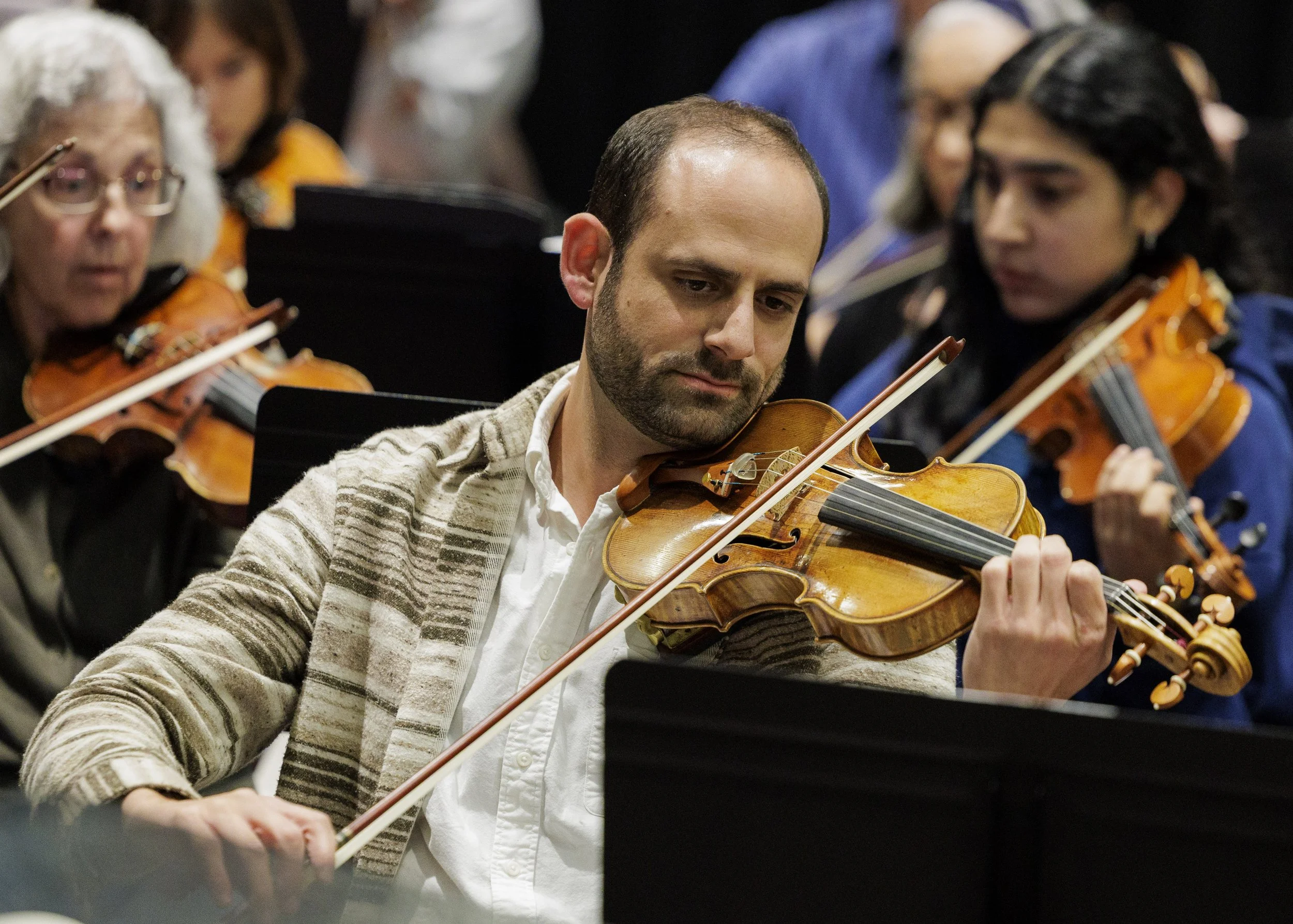 A group of musicians playing violins in an orchestra, with a focus on a man in the foreground leading the section with a serious expression.