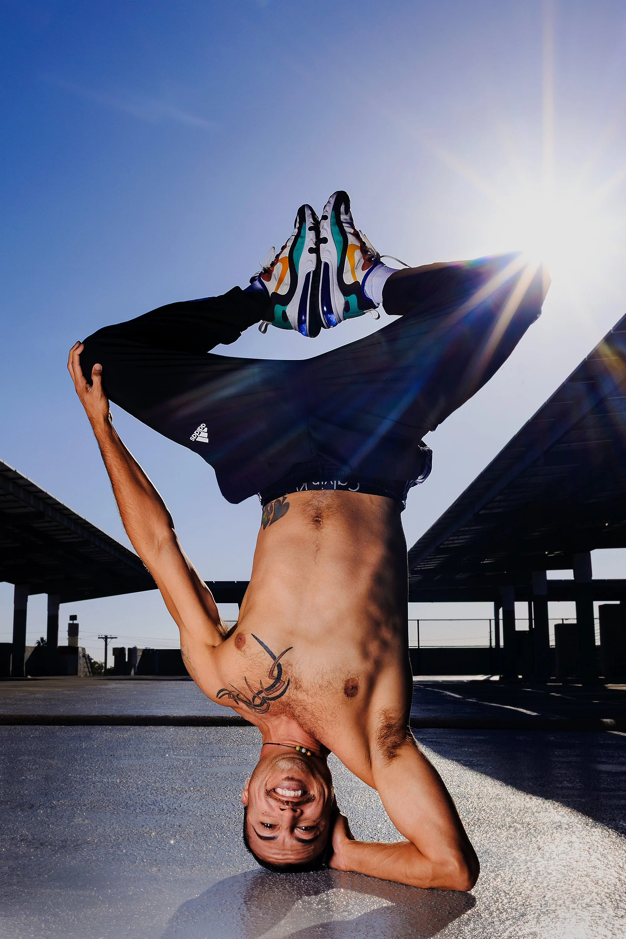 A shirtless man doing a handstand on a wet pavement under a bright sun, with his legs bent and feet touching, wearing colorful sneakers and black pants, in an outdoor parking lot.